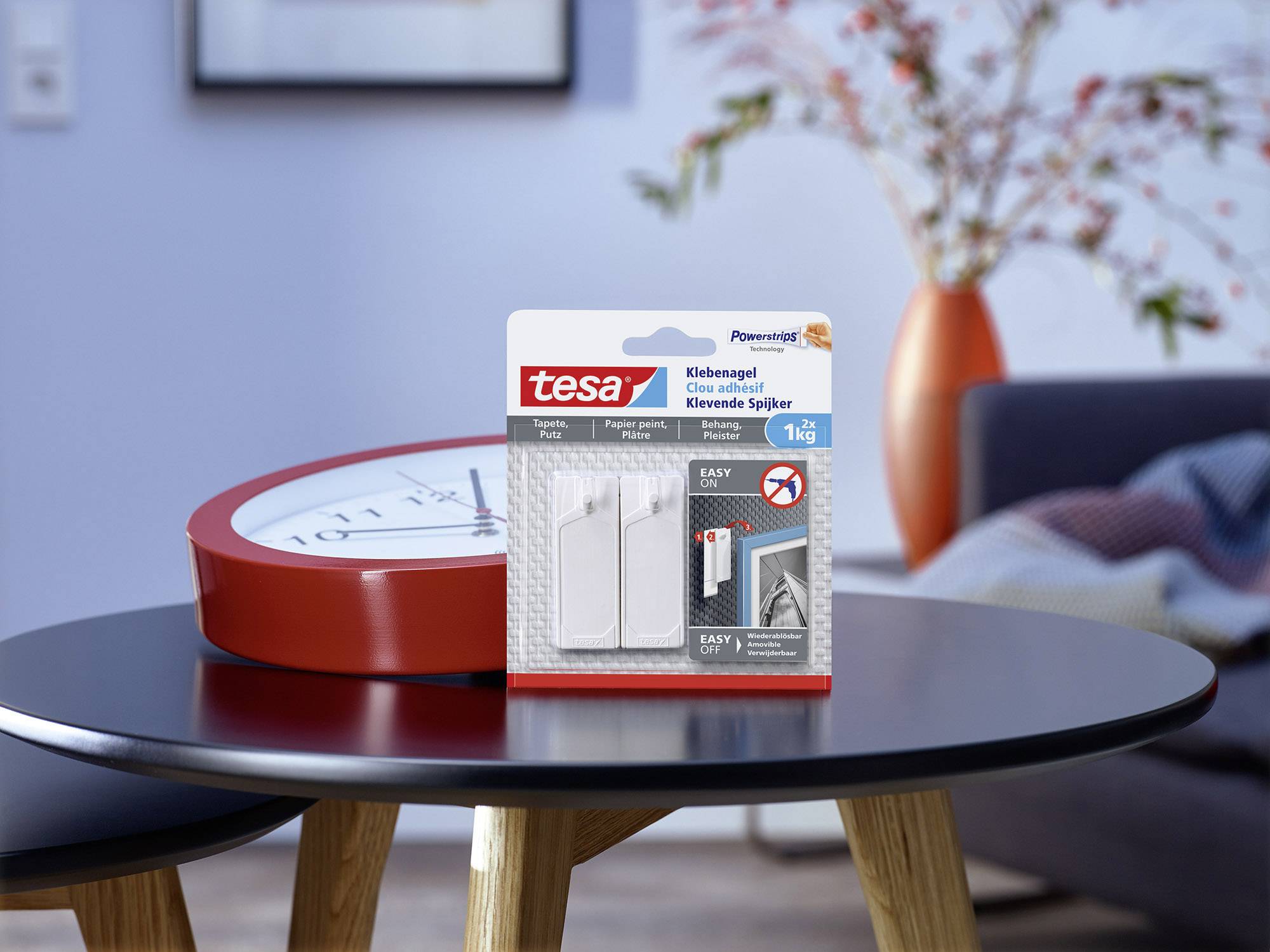 tesa Klebenagel-Packung mit 1 kg Tragfähigkeit auf einem Tisch in einem Wohnzimmer mit roter Uhr und bunter Vase im Hintergrund.