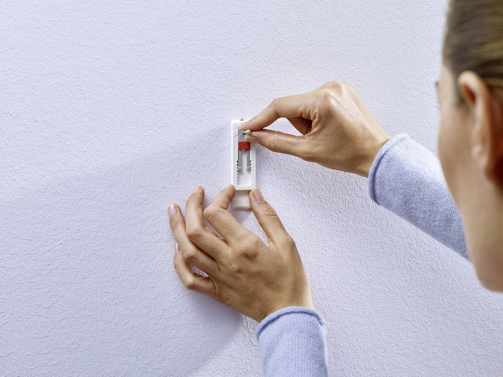 Eine Person montiert mit beiden Händen eine kleine rote und weiße Wasserwaage an einer hellen Wand.