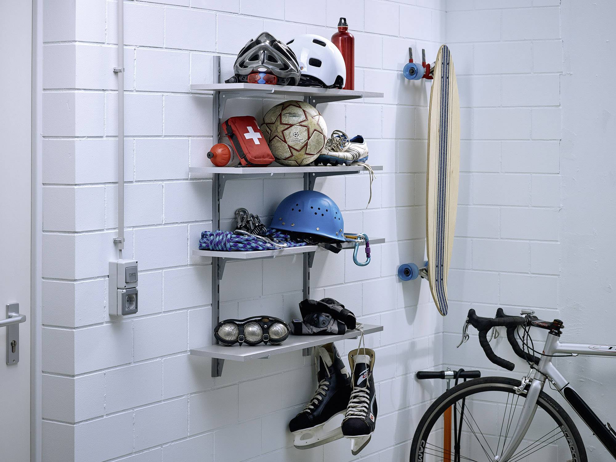 'Garagenwand mit Regalen voller Sportausrüstung: Helme, Fußbälle, Inlineskates, Erste-Hilfe-Set; daneben ein Fahrrad und ein Longboard.'