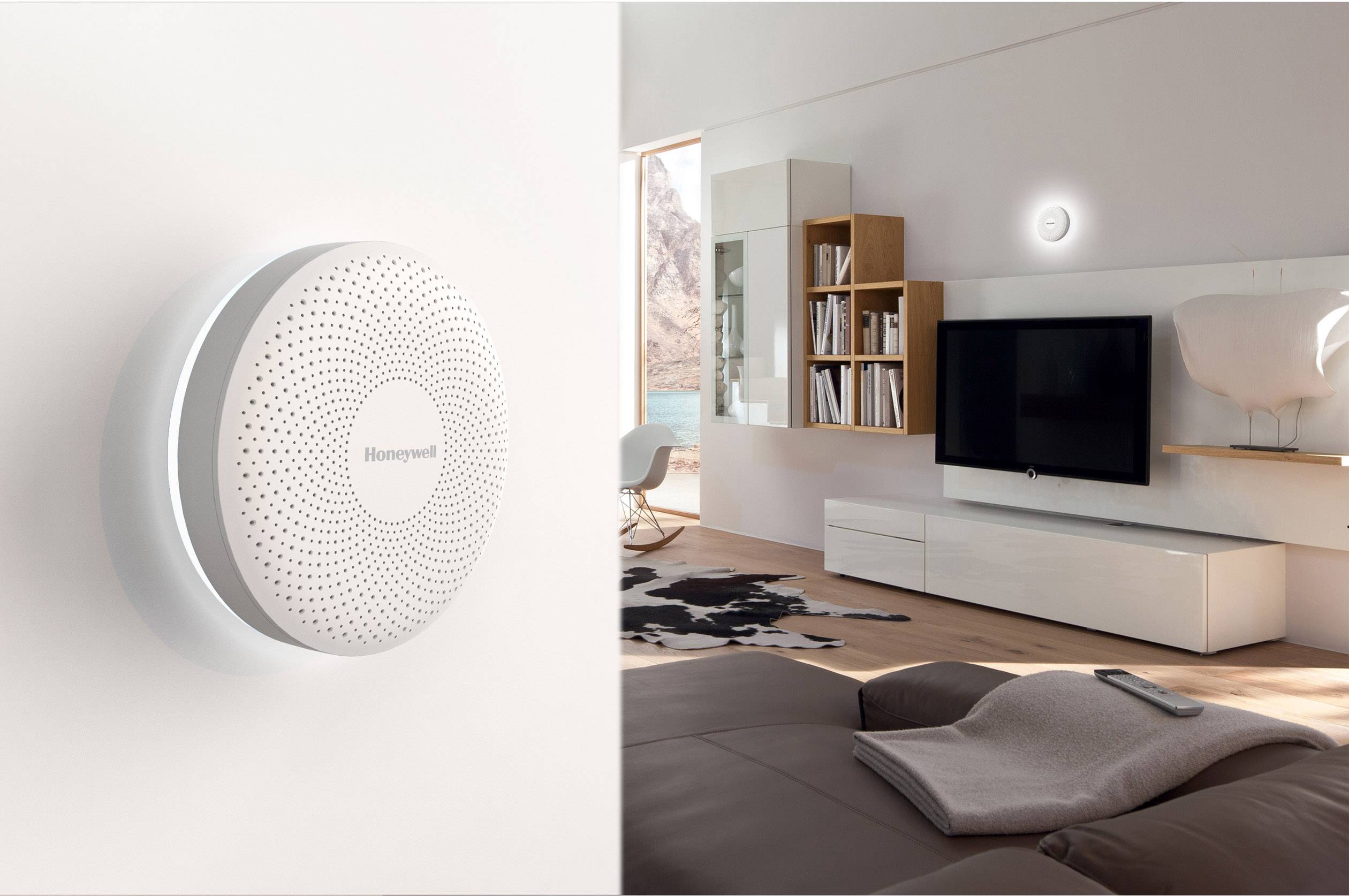 Ein Rauchmelder ist an der Wand eines modernen, hellen Wohnzimmers montiert. Im Hintergrund stehen ein Fernseher und Regale.