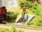 Ein rotes Eisenbahnsignal steht neben Bahngleisen in einer Miniaturlandschaft, umgeben von grüner Vegetation und einem Backsteinhäuschen.