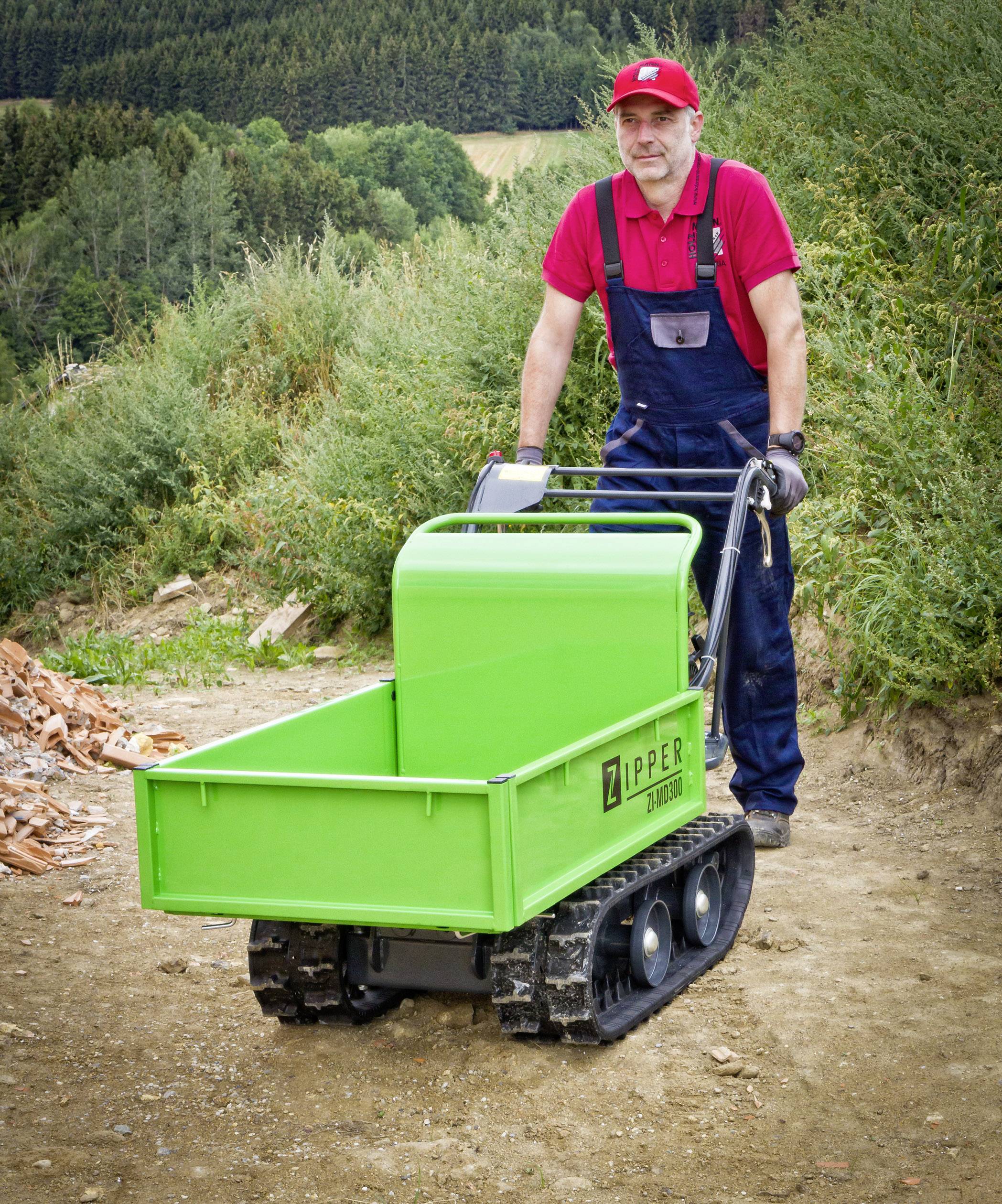 Ein Mann schiebt auf einem Waldweg eine grüne motorisierte Schubkarre. Im Hintergrund sind Bäume und Sträucher zu sehen.