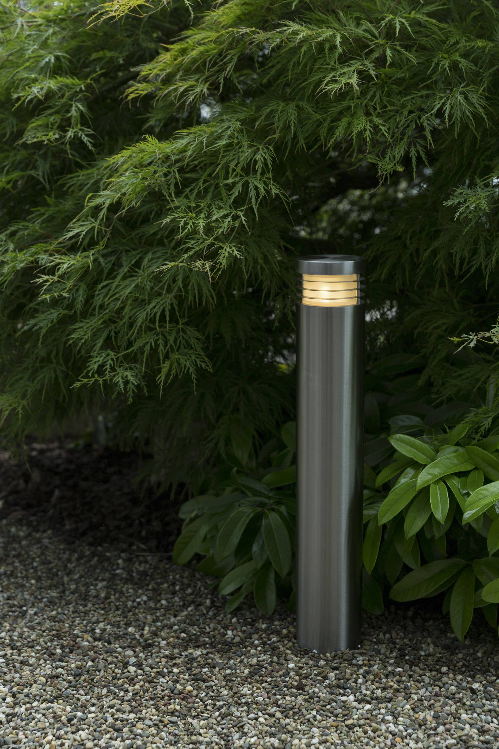 Eine moderne, zylindrische Außenlampe leuchtet dezent im Garten, umgeben von grünem Laub und Kies, schafft eine ruhige Atmosphäre.