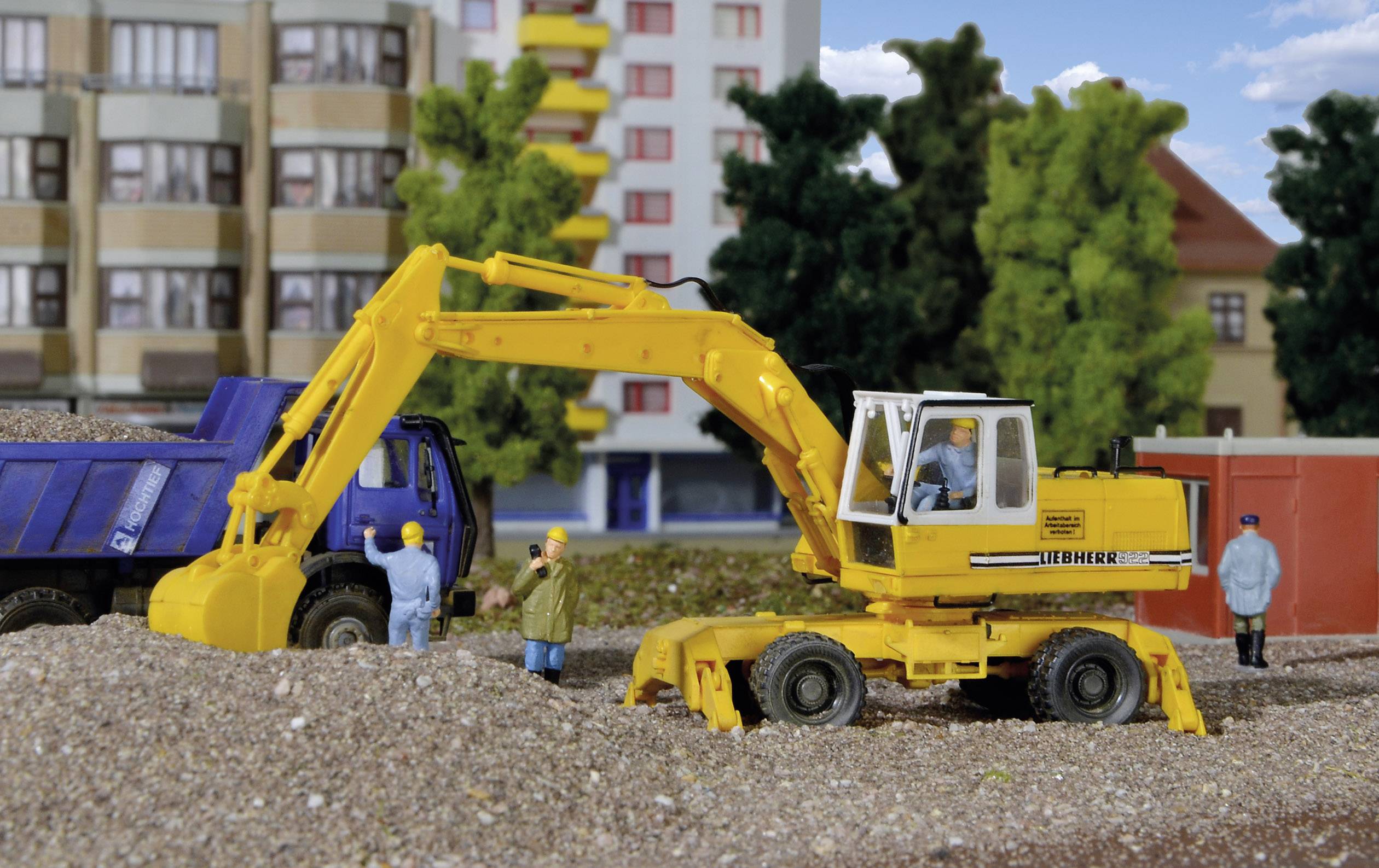 Ein gelber Bagger lädt Erde auf einen blauen Lastwagen. Zwei Bauarbeiter in Helm beobachten. Im Hintergrund sind Gebäude und Bäume zu sehen.