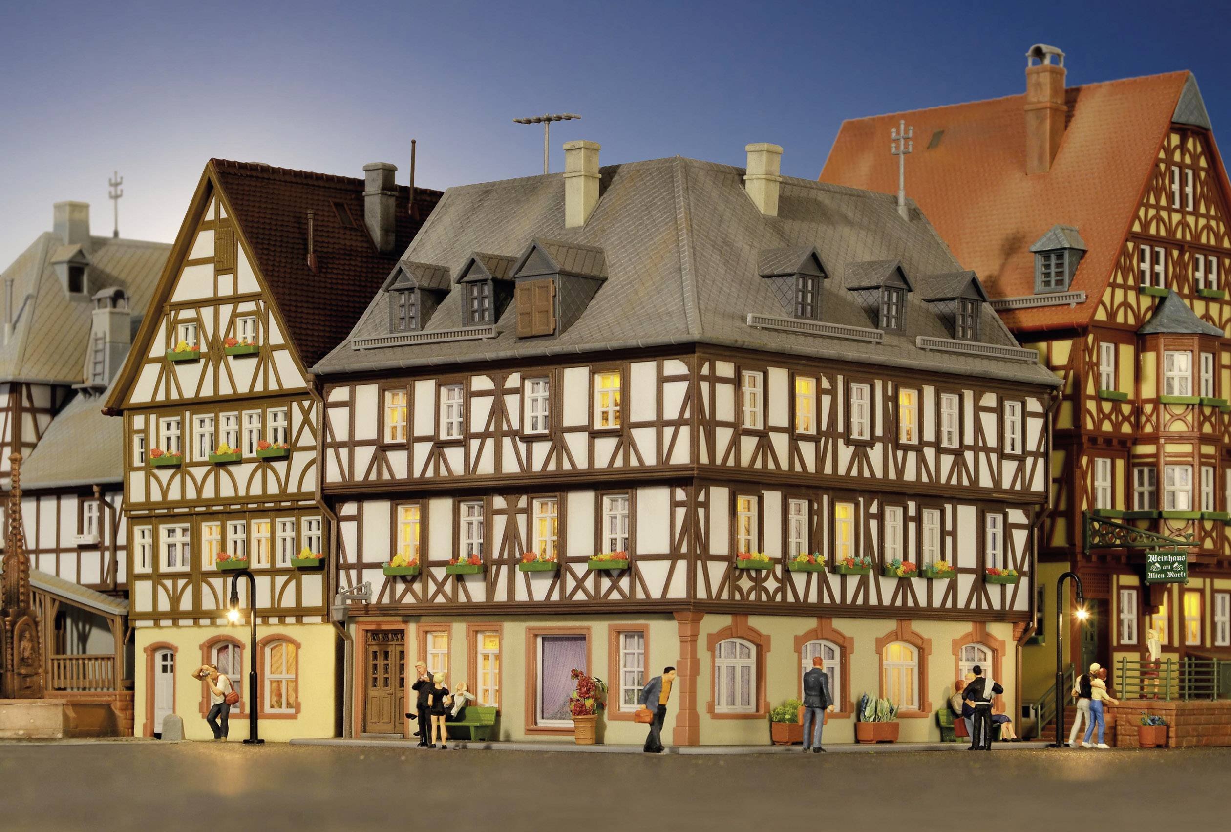 Fachwerkhäuser mit leuchtenden Fenstern in einer Miniaturstadt bei Dämmerung, Menschen gehen und unterhalten sich auf der Straße.