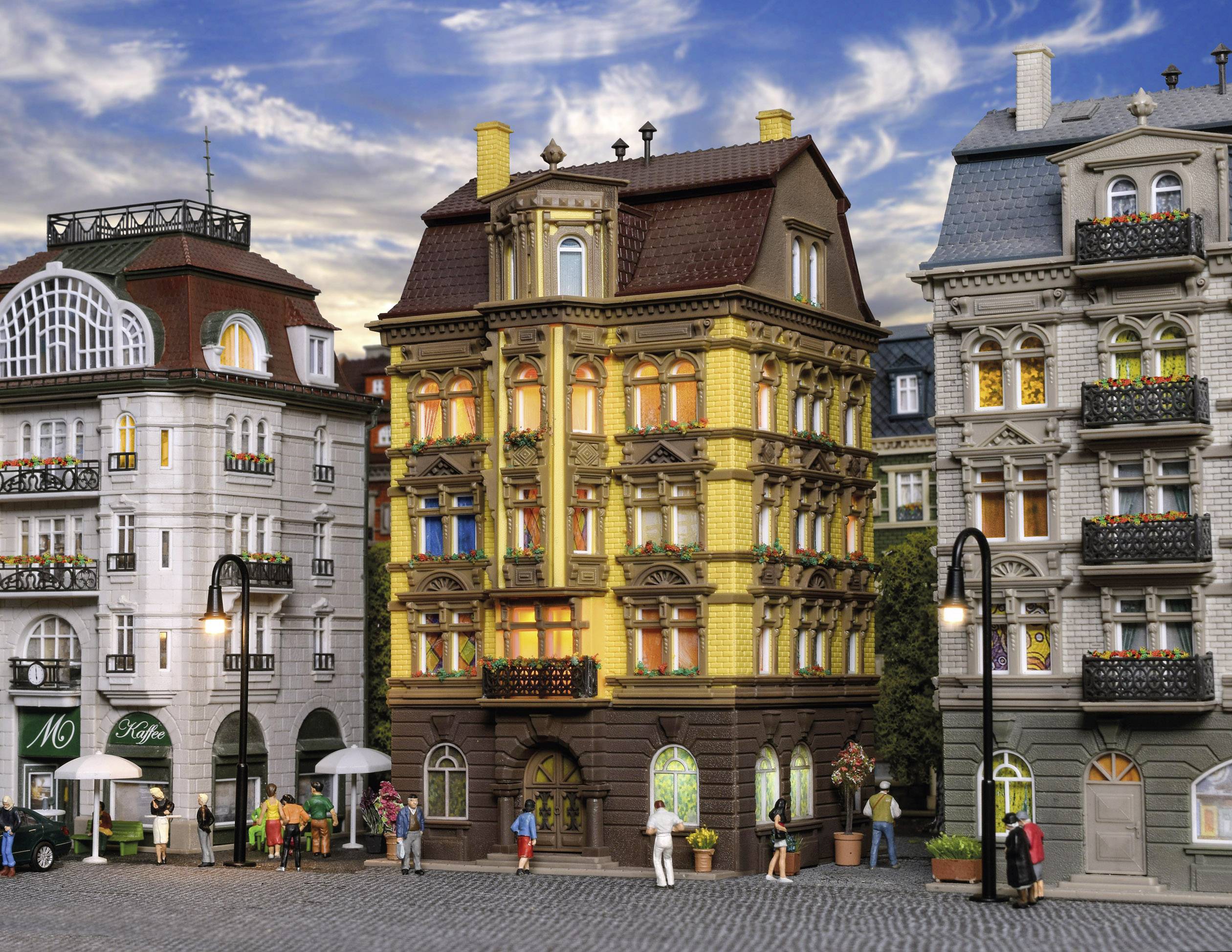 Mehrere farbenfrohe, detaillierte Modelle von Stadthäusern, umgeben von Miniaturfiguren, die das Stadtleben darstellen; blauer Himmel im Hintergrund.