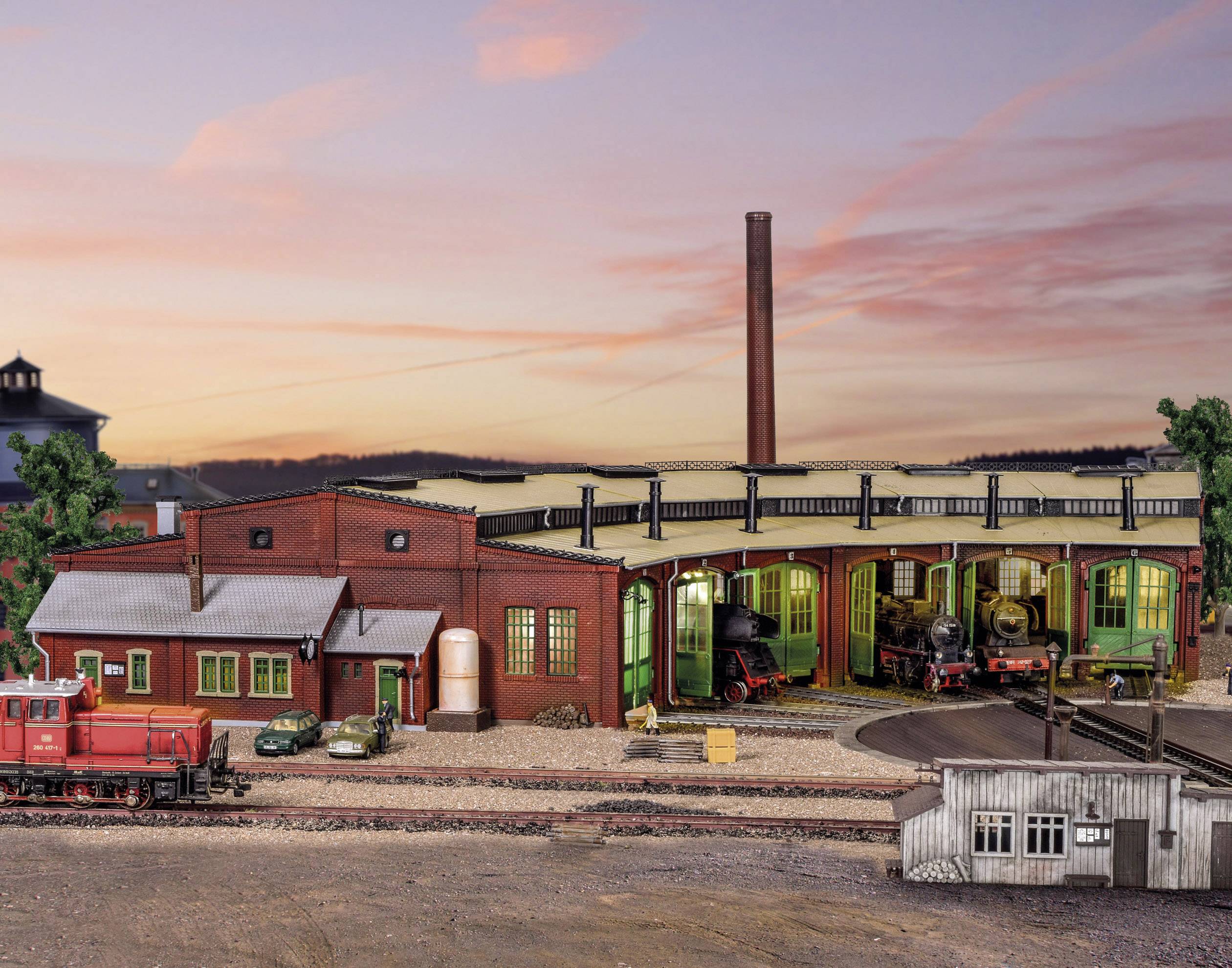 Eine Modelleisenbahn-Werkstatt mit Zügen, umgeben von Gebäuden und Landschaft im Miniaturformat bei Sonnenuntergang.