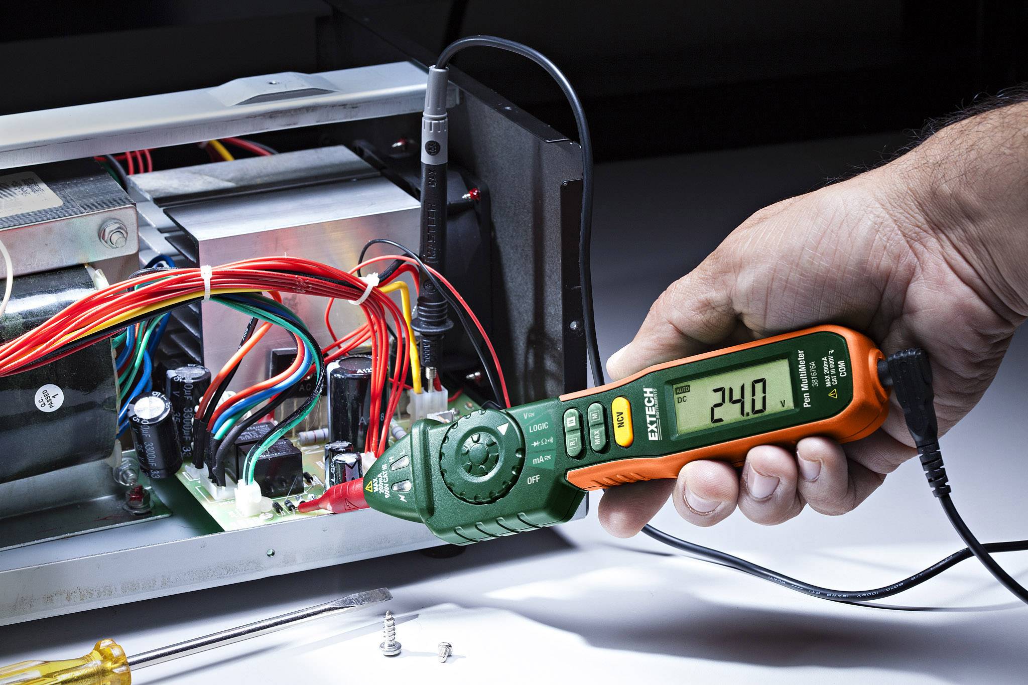 Eine Person hält ein Multimeter an eine geöffnete elektrische Schaltung, das Display zeigt '24.0'. Kabel und Komponenten sind sichtbar.