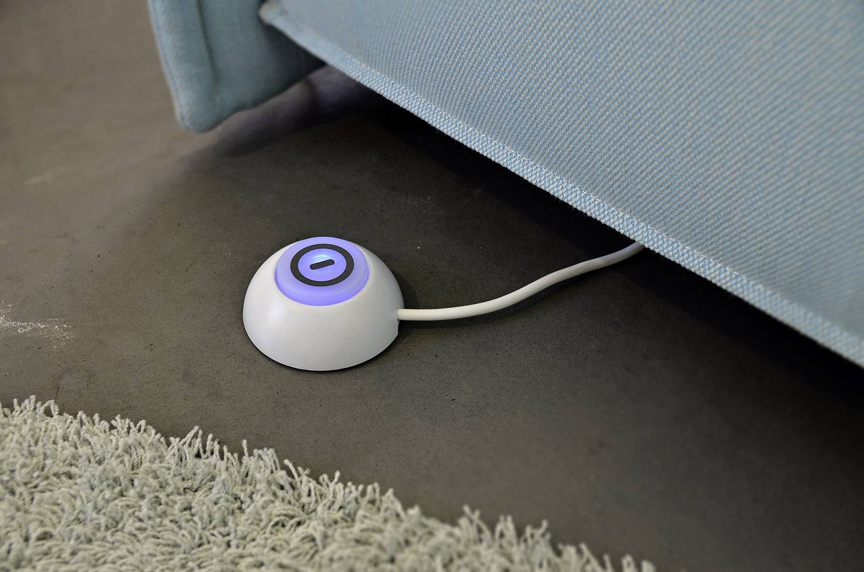 Ein runder, weißer Fußschalter mit blauem Leuchtring unter einem Sofa auf einem Betonboden neben einem Teppich.