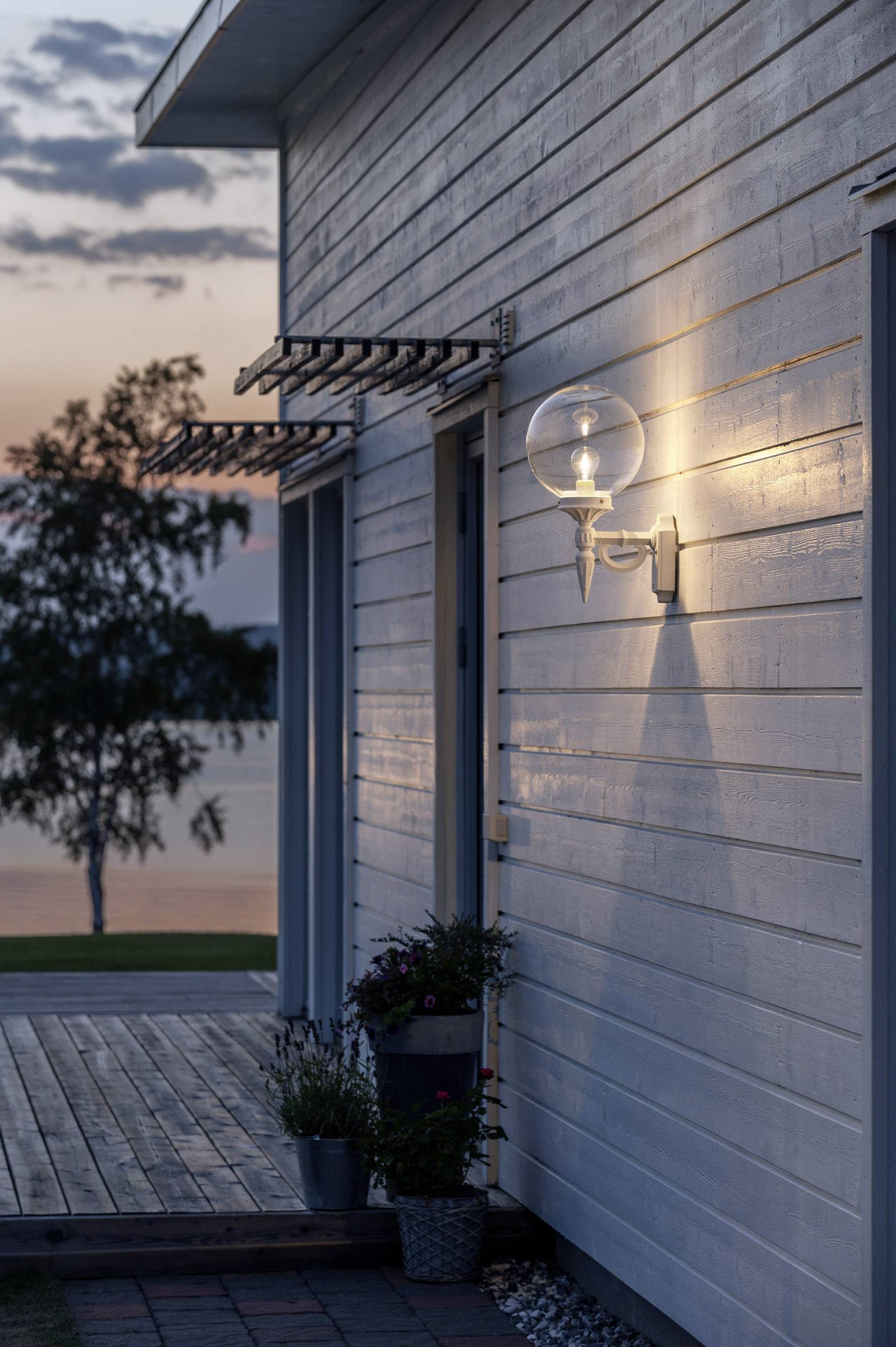 Eine moderne Holzhauswand bei Sonnenuntergang, beleuchtet von einer runden Außenlampe. Im Hintergrund ein Baum am Ufer eines Sees.