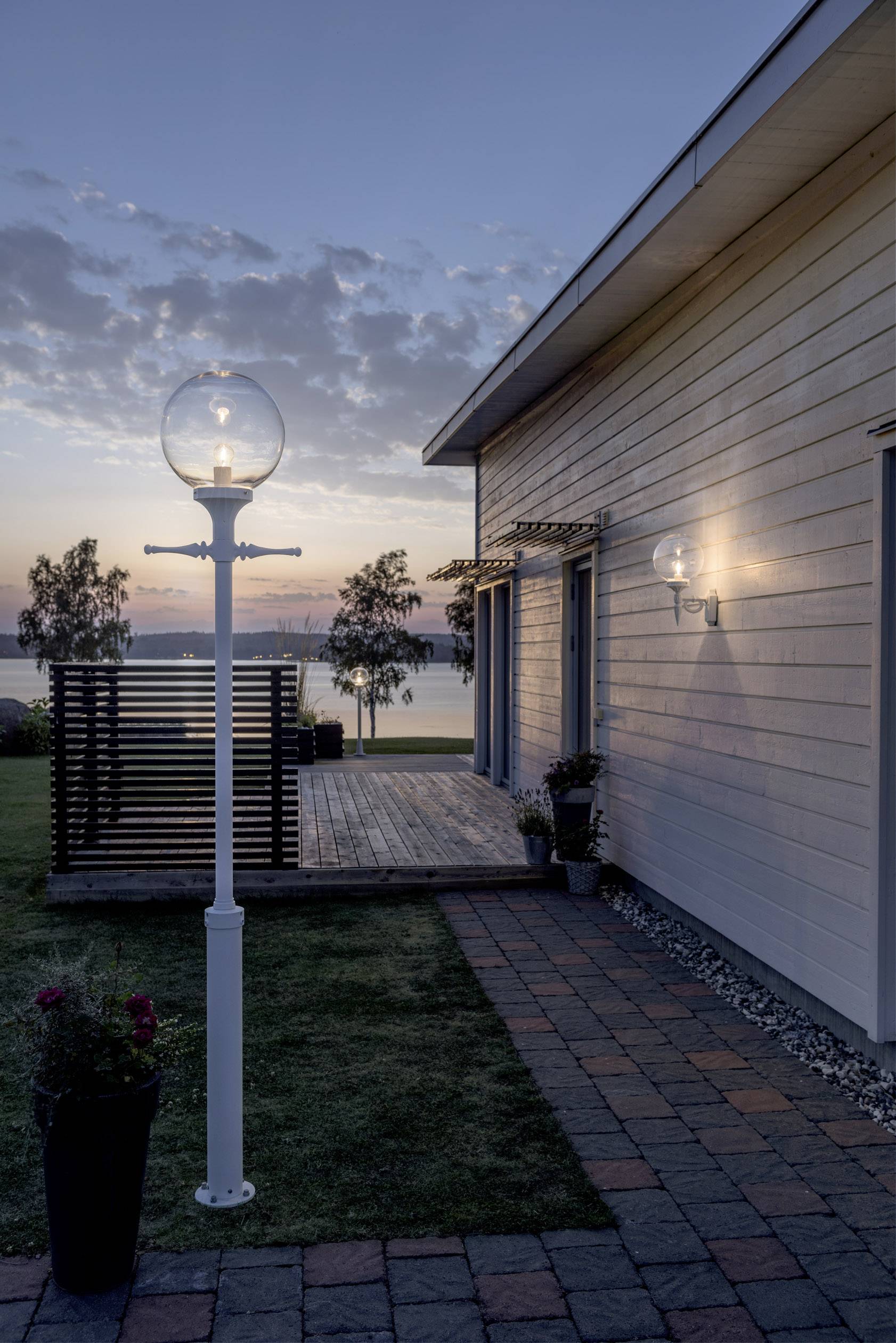 Ein modernes Haus bei Sonnenuntergang, mit beleuchteter Außenlampe und Seeblick im Hintergrund. Gartenelemente und Pflanze im Vordergrund.