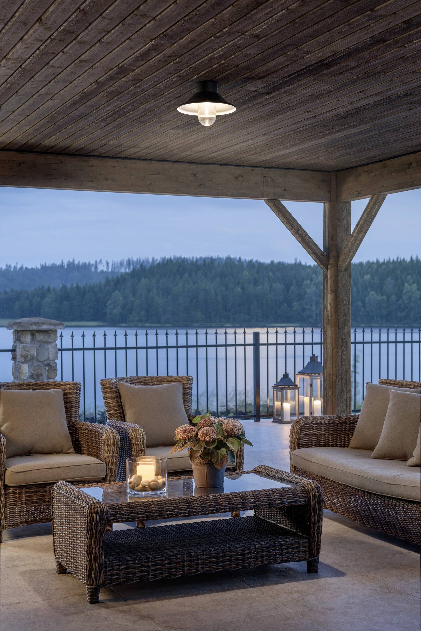 Überdachte Terrasse mit Rattanmöbeln und Blick auf einen See bei Sonnenuntergang. Laternen und Kerzen sorgen für stimmungsvolle Beleuchtung.