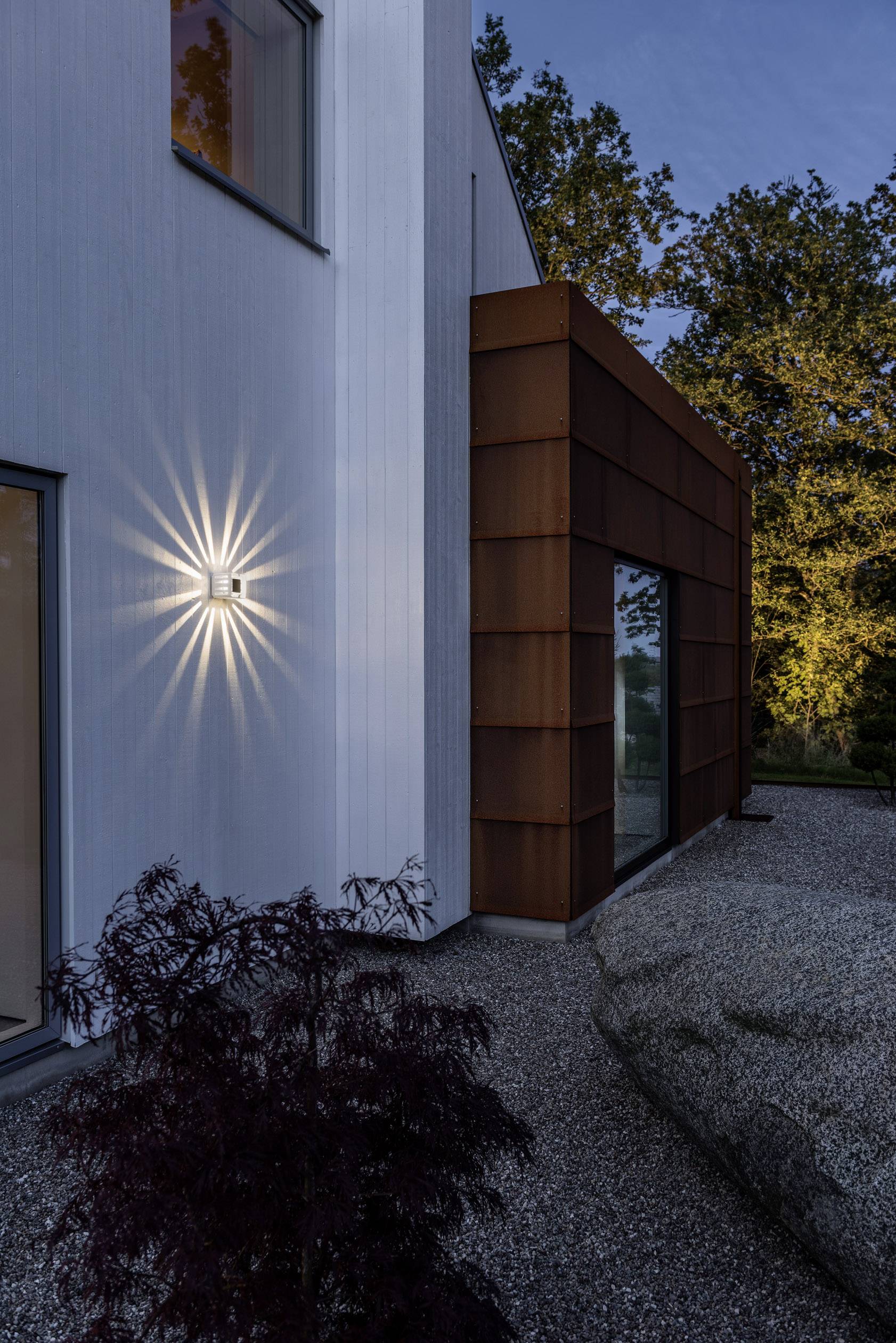 Moderne Architektur: Eine weiß gestrichene Hauswand mit einer leuchtenden Außenlampe. Rostrote Verkleidung grenzt daran, umgeben von Bäumen.