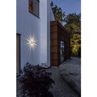 Moderne Architektur: Eine weiß gestrichene Hauswand mit einer leuchtenden Außenlampe. Rostrote Verkleidung grenzt daran, umgeben von Bäumen.