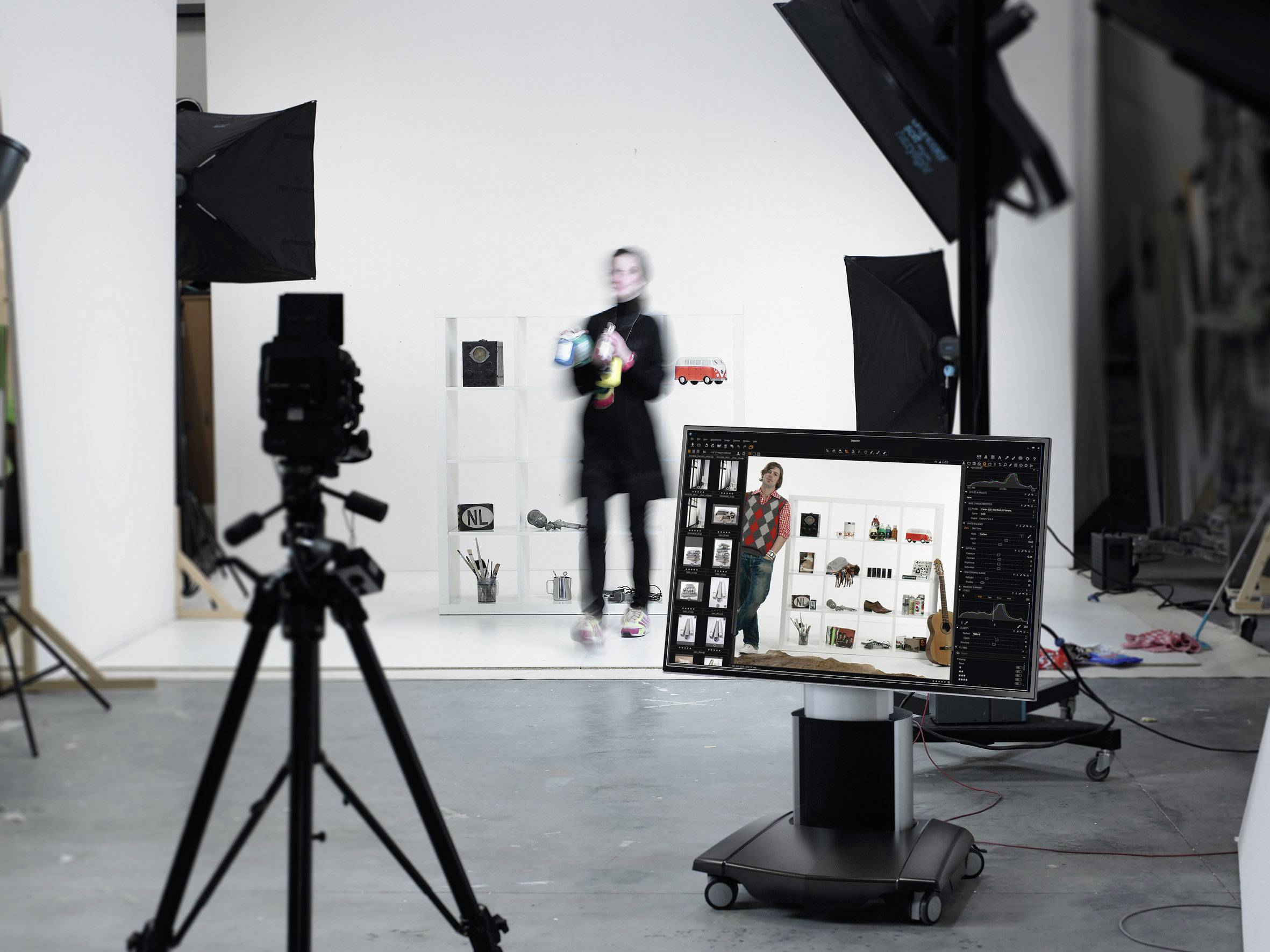 Eine Fotostudioaufnahme: Eine Person steht vor einem Regal mit Kameras und Accessoires. Im Vordergrund sind Kameras und ein Computerbildschirm zu sehen.