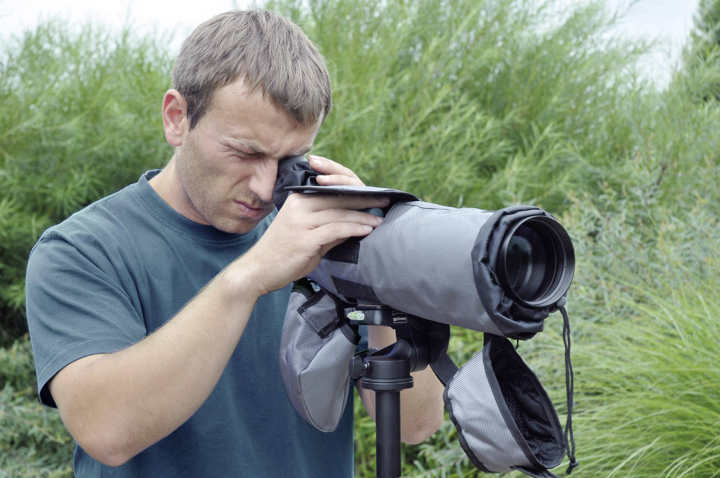 Ein Mann betrachtet durch ein großes Teleskop auf einem Stativ im Freien, umgeben von grünem Gebüsch.