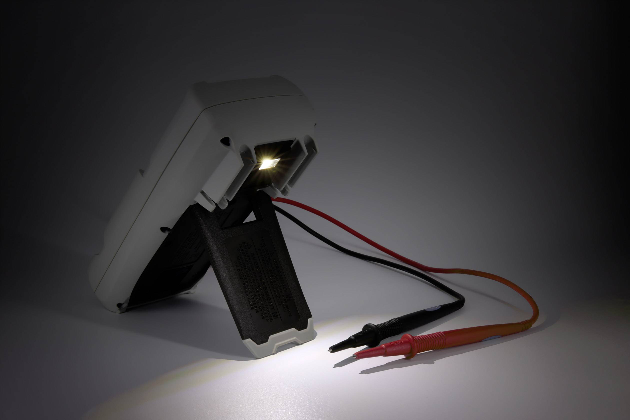 Ein Multimeter steht schräg auf einem Ständer, beleuchtet durch eine kleine Lampe. Ein schwarzes und ein rotes Kabel hängen herab.