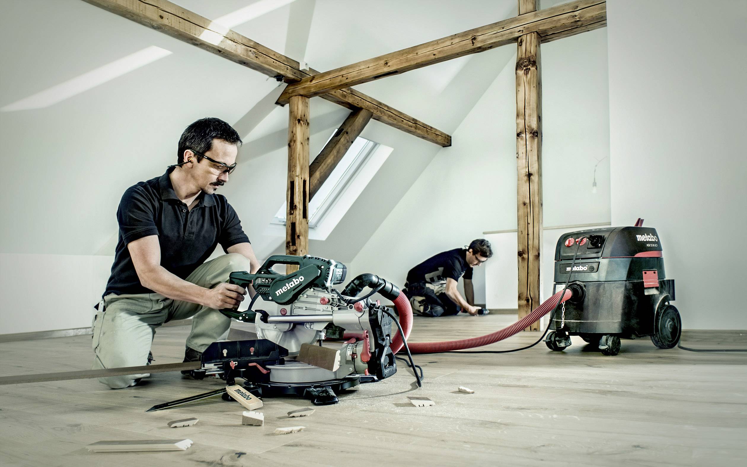 Zwei Handwerker arbeiten auf einem Dachboden. Einer benutzt eine Kappsäge, der andere verlegt Holzboden. Elektrische Geräte sind sichtbar.