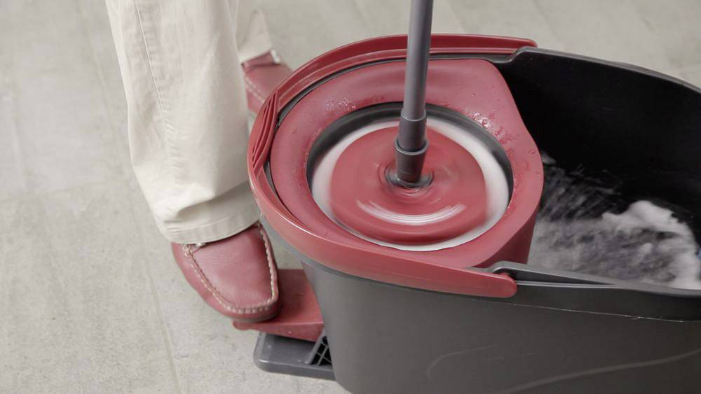 Eine Person bedient einen Mopp-Eimer mit Fußpedal, um Wasser aus dem Moppkopf auszuwringen. Der Eimer ist teilweise gefüllt.