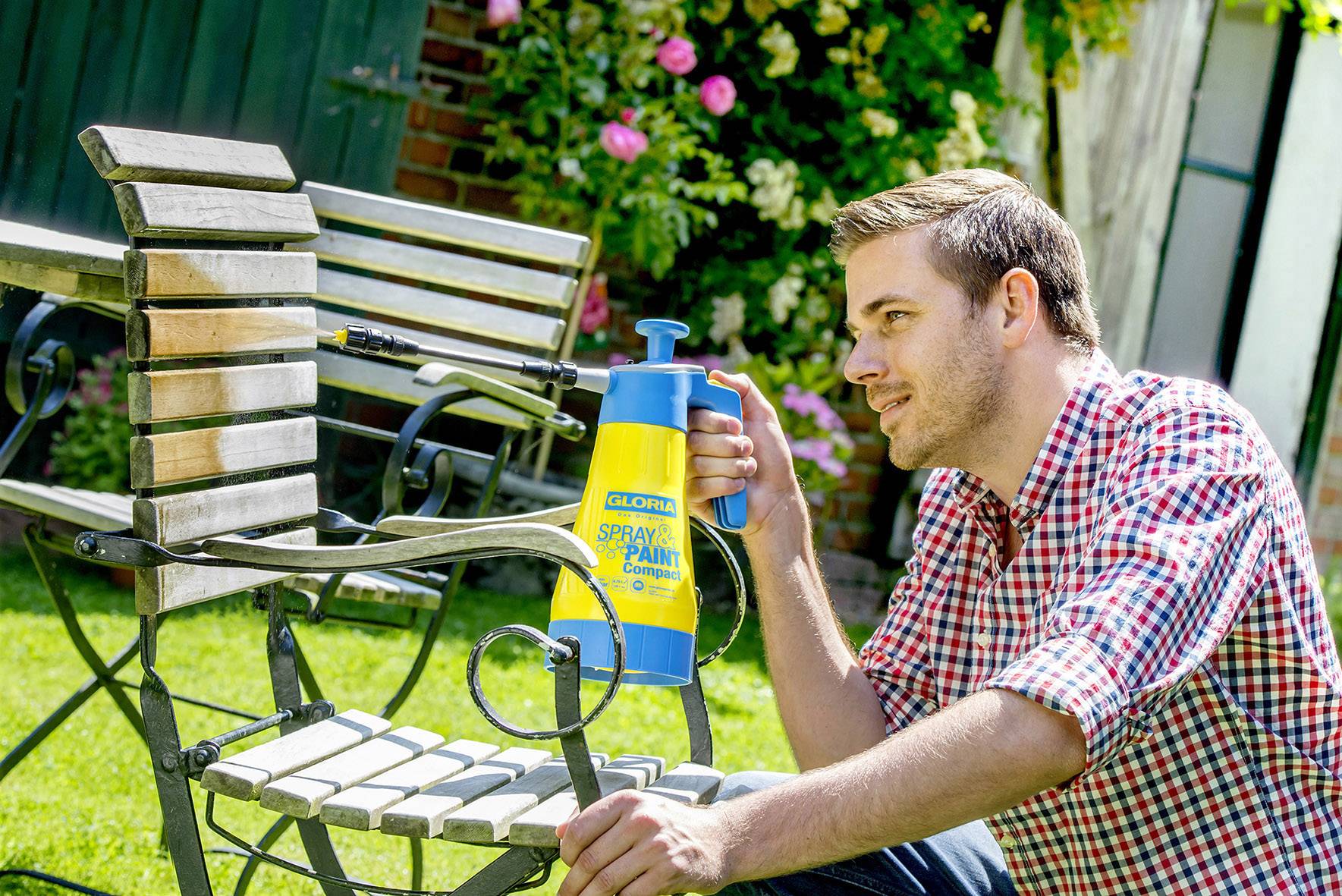 Ein Mann besprüht im Garten eine Metallbank mit einer gelben Sprühflasche. Im Hintergrund sind Blumenbeete zu sehen.