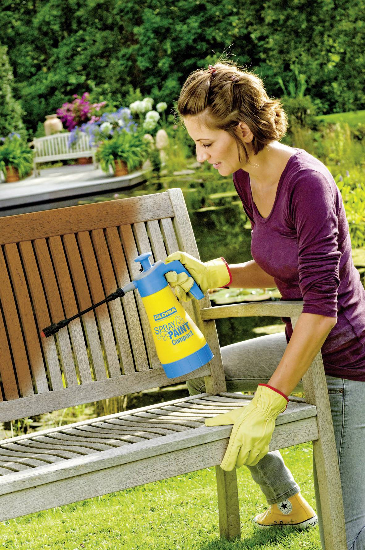 Eine Person reinigt eine Holzbank mit einem Sprühreiniger im Garten. Im Hintergrund sind Pflanzen und ein Teich sichtbar.