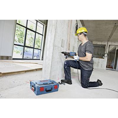 Ein Bauarbeiter kniet auf dem Boden eines Gebäuderohbaus und bohrt mit einem Elektrowerkzeug Löcher in eine Betonwand.
