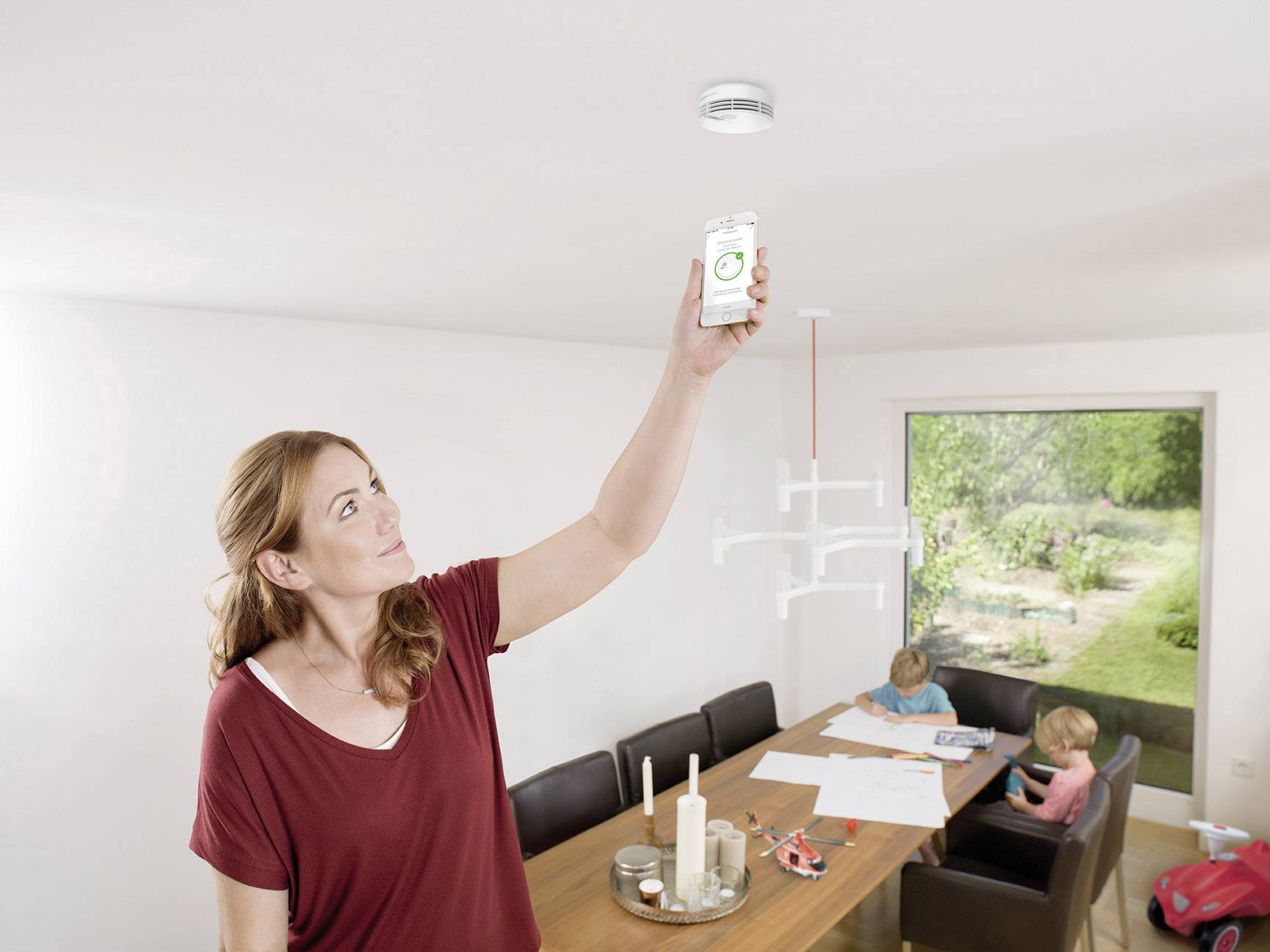 Eine Frau überprüft Rauchmelder mit Smartphone in hell beleuchtetem Esszimmer, während zwei Kinder am Tisch malen.