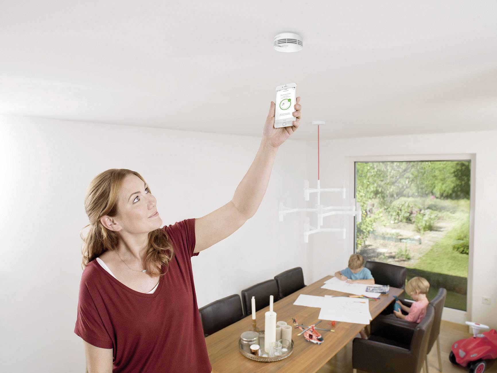 Eine Frau testet mit ihrem Smartphone einen Rauchmelder an der Zimmerdecke, zwei Kinder malen am Tisch im Hintergrund.