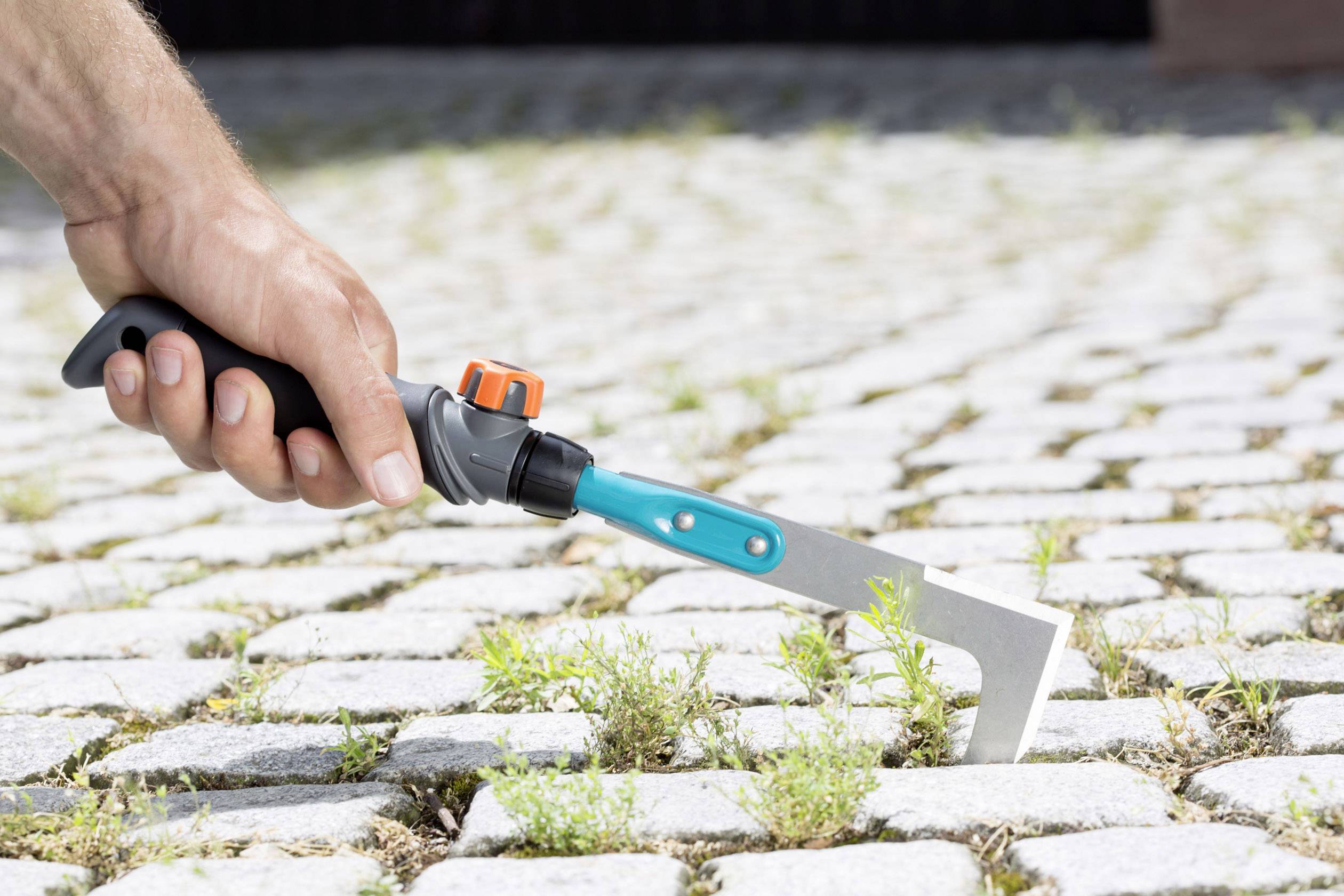 Eine Hand benutzt ein Fugenkratzer, um Unkraut zwischen Pflastersteinen zu entfernen.