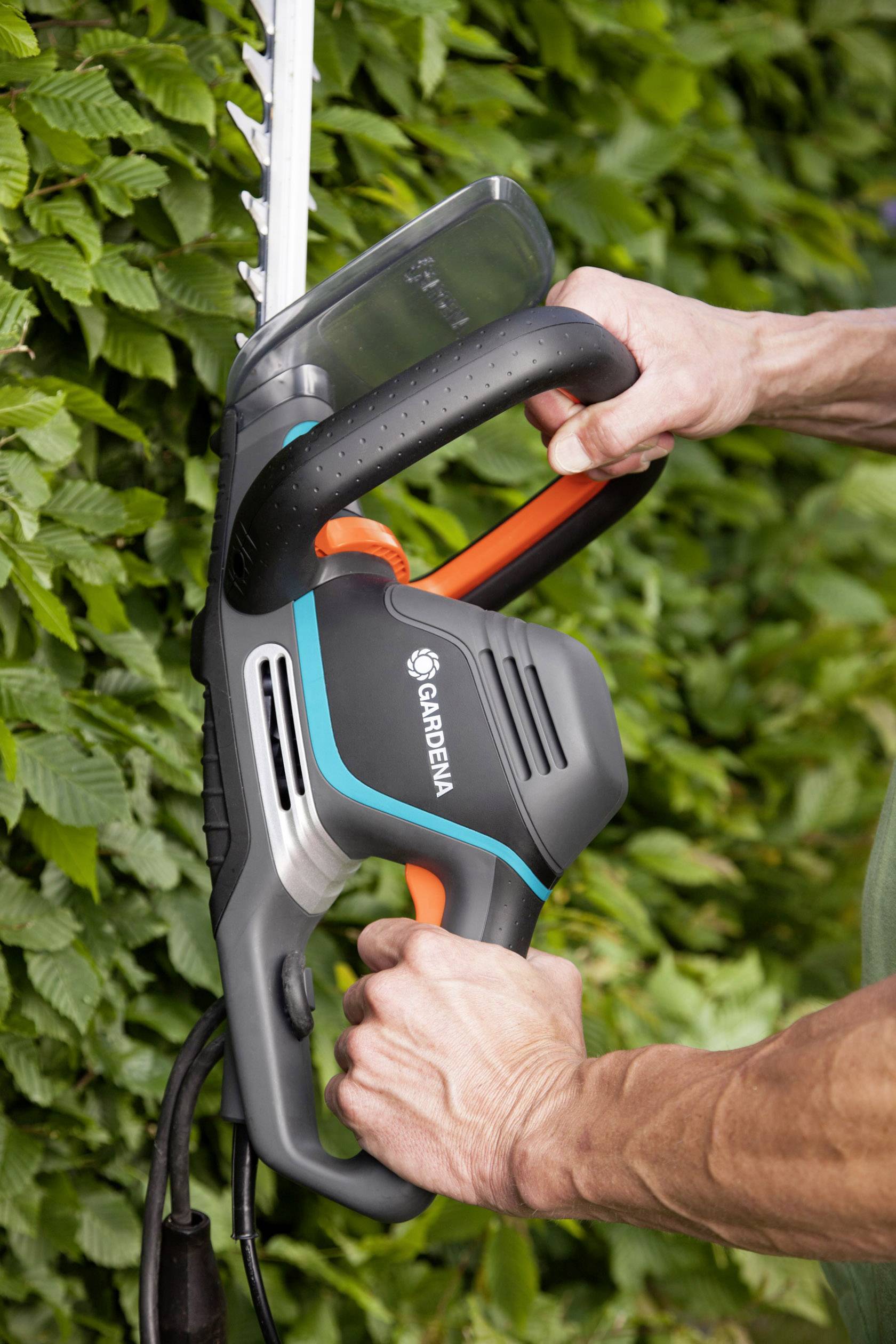 Eine Person schneidet mit einem elektrischen Heckenschneider von Gardena eine grüne Hecke. Hände halten den Griff des Werkzeugs fest.