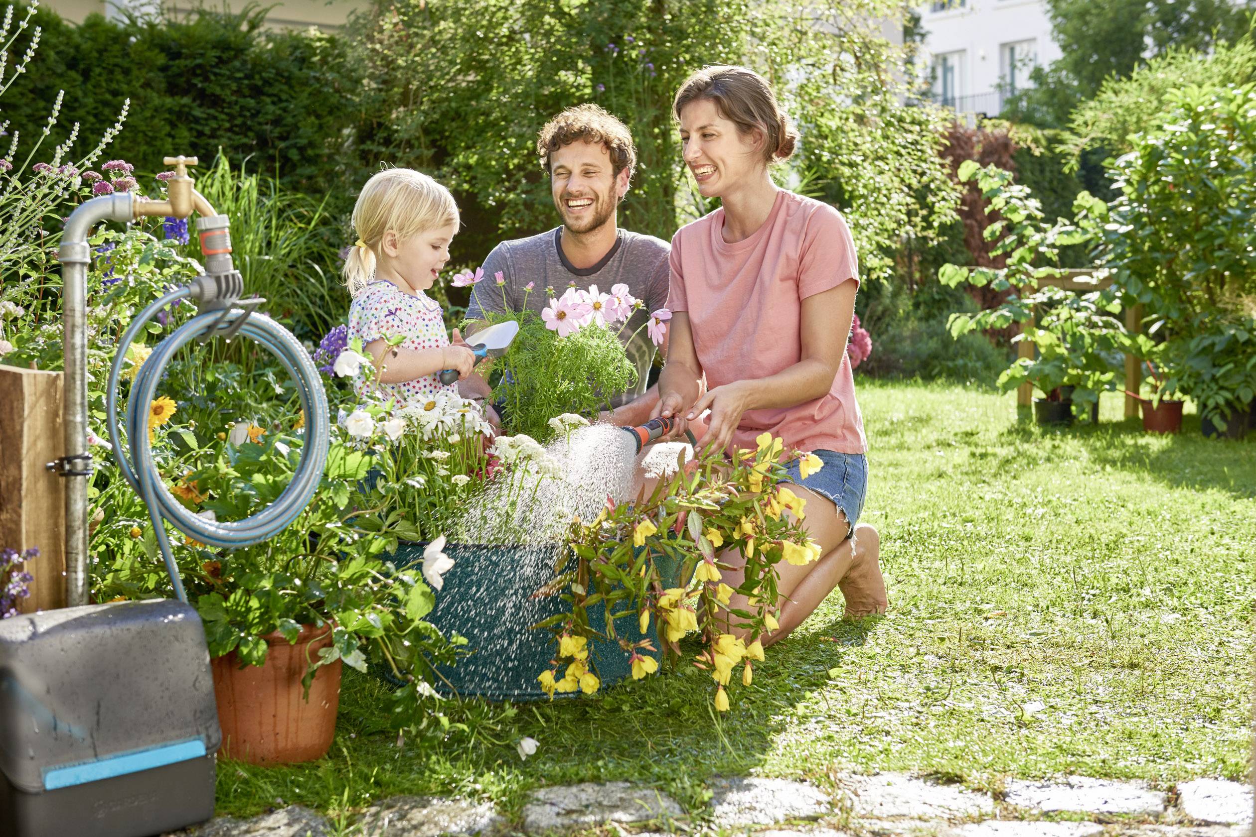 Eine Familie sitzt im Garten und pflanzt Blumen. Ein Mann, eine Frau und ein Kind lächeln und gießen gemeinsam Pflanzen.