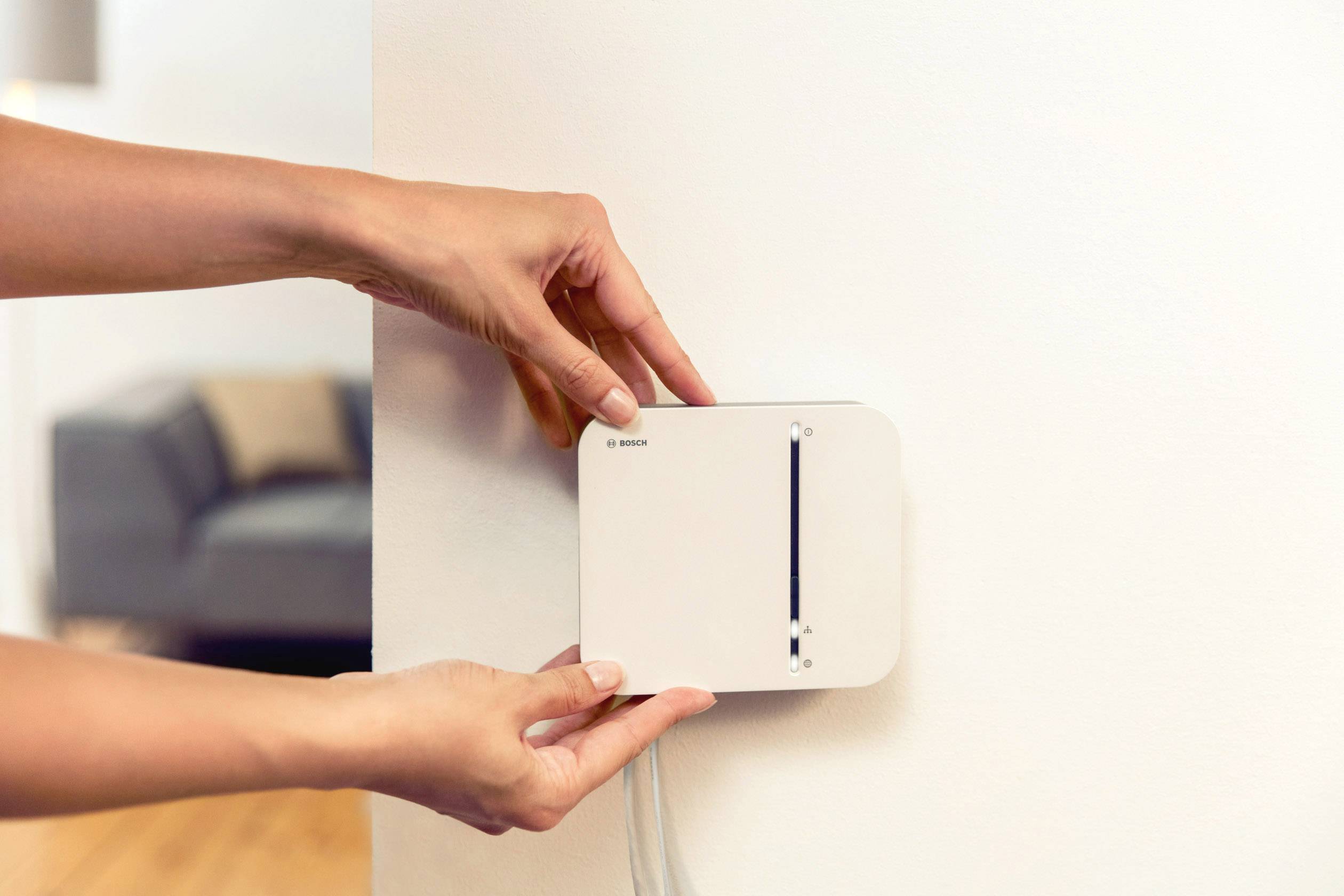 Eine Person montiert ein weißes, rechteckiges Gerät an einer Wand. Im Hintergrund unscharf ist ein Wohnzimmer zu sehen.