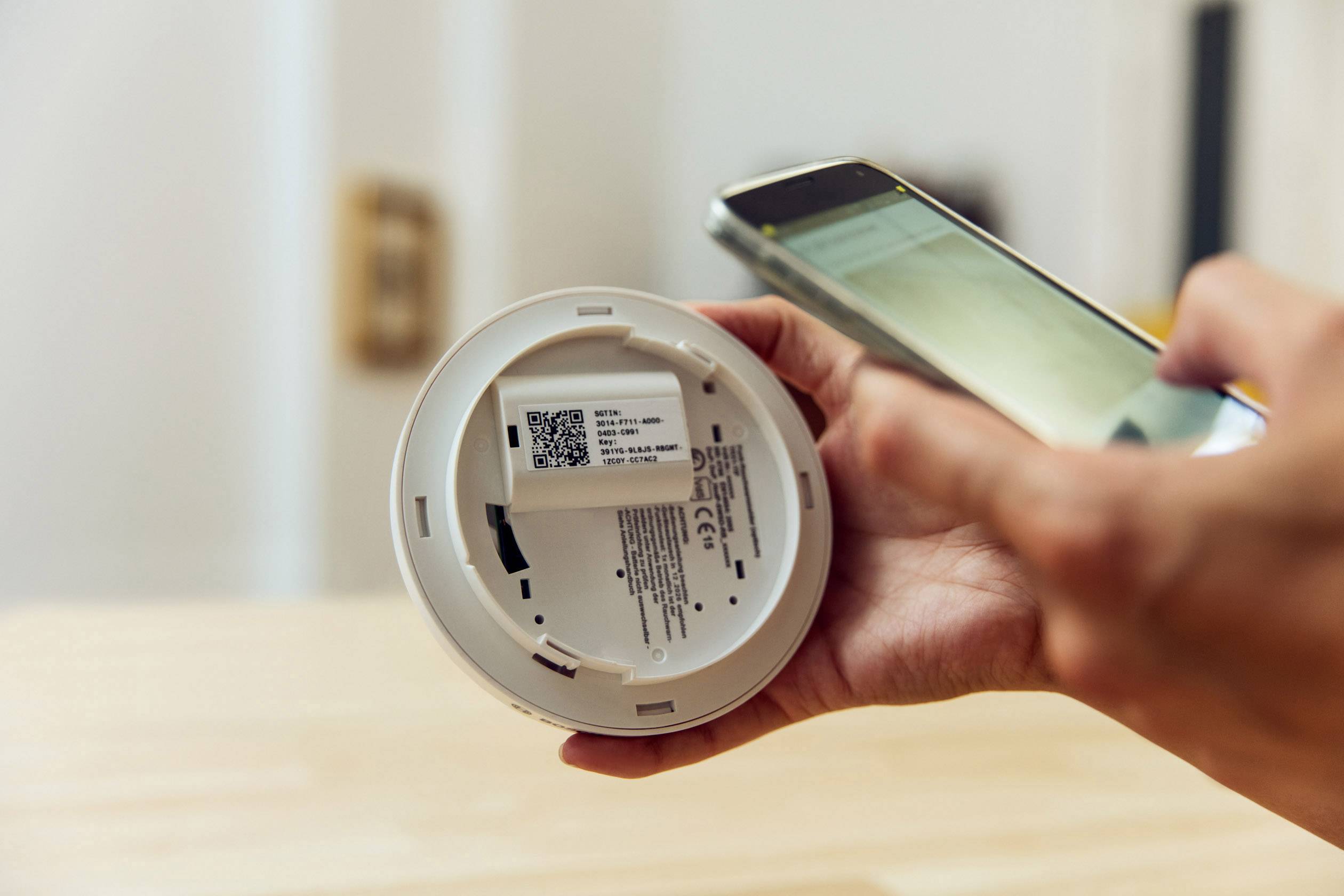 Eine Person scannt den QR-Code auf der Rückseite eines Rauchmelders mit einem Smartphone. Im Hintergrund sind verschwommene Türrahmen.
