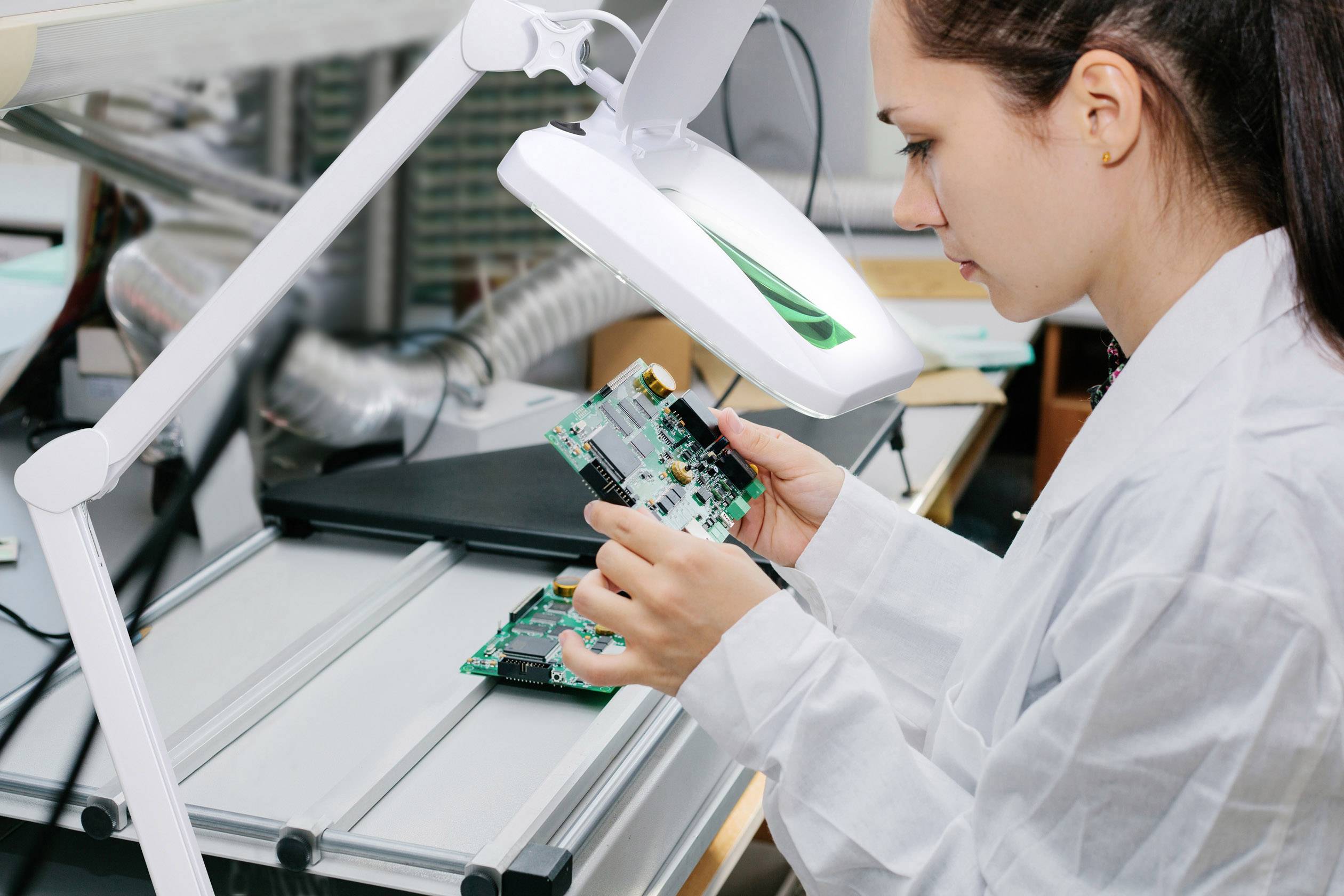 Ingenieurin in weißem Kittel inspiziert eine Leiterplatte unter einer Vergrößerungslampe in einem Labor.