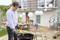 Familie bei Grillfeier im Garten. Ein Mann zündet Kohlen im Grill an, während eine Frau und ein Kind im Hintergrund auf der Terrasse stehen.