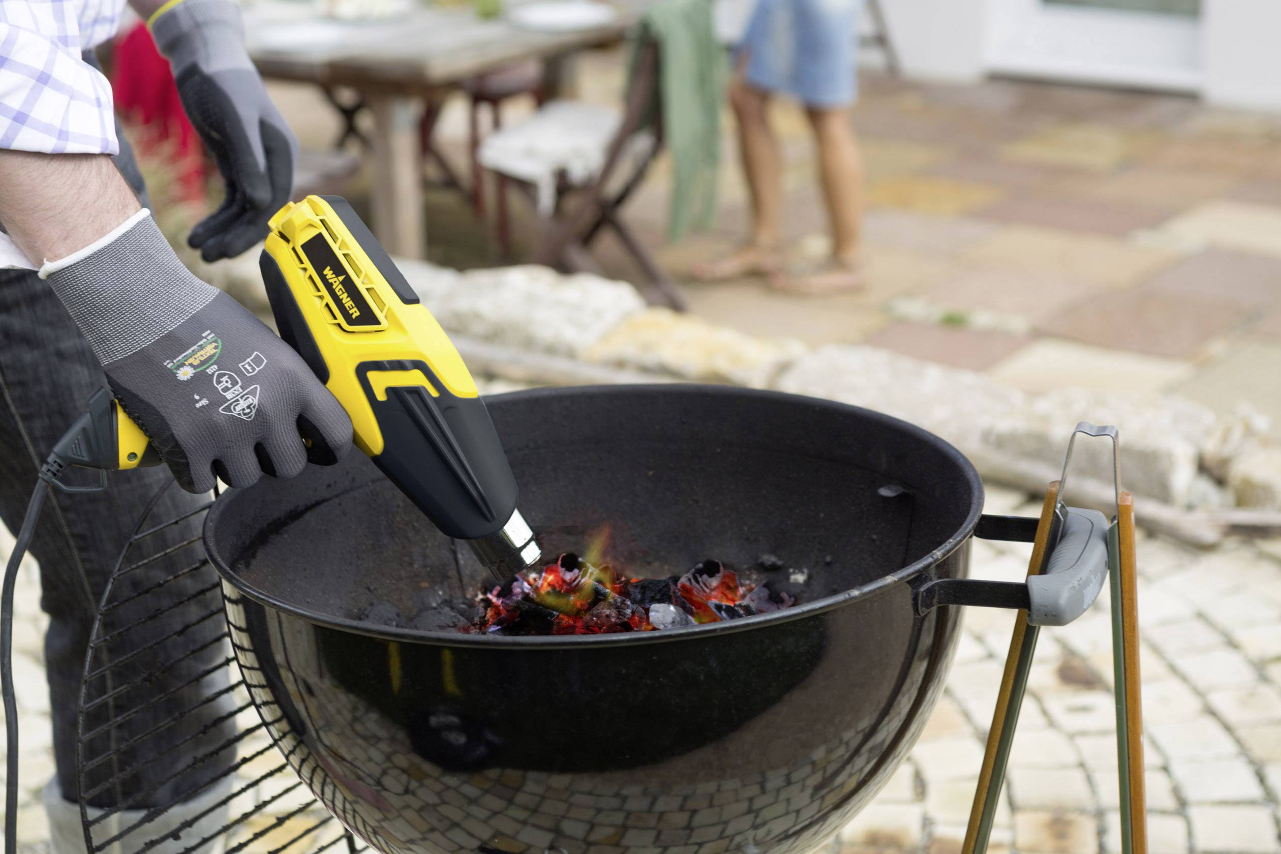 Ein Grill wird mit einem gelben Heißluftgebläse angezündet. Im Hintergrund sind unscharf Personen und eine Tischlandschaft zu sehen.