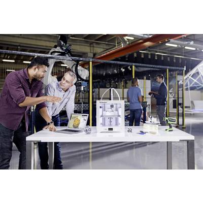 Zwei Männer diskutieren in einer Werkstatt über einen 3D-Drucker auf einem Tisch. Im Hintergrund arbeiten weitere Personen.