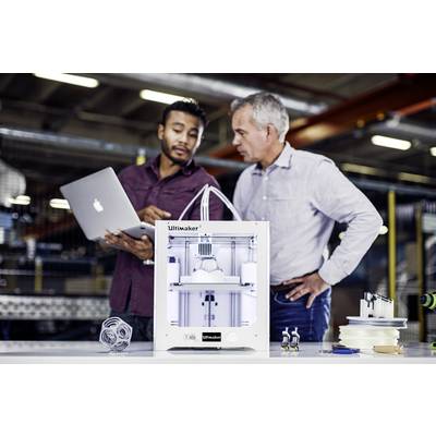 Zwei Männer betrachten einen 3D-Drucker auf einem Tisch. Einer hält einen Laptop, beide scheinen in einer Fabrikumgebung zu arbeiten.
