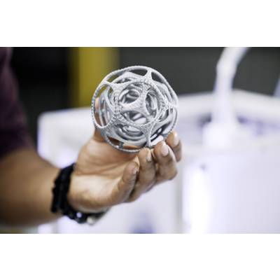 Eine Hand hält ein komplex modelliertes, graues 3D-gedrucktes Objekt in Kugelform, unscharfer Hintergrund mit 3D-Drucker.