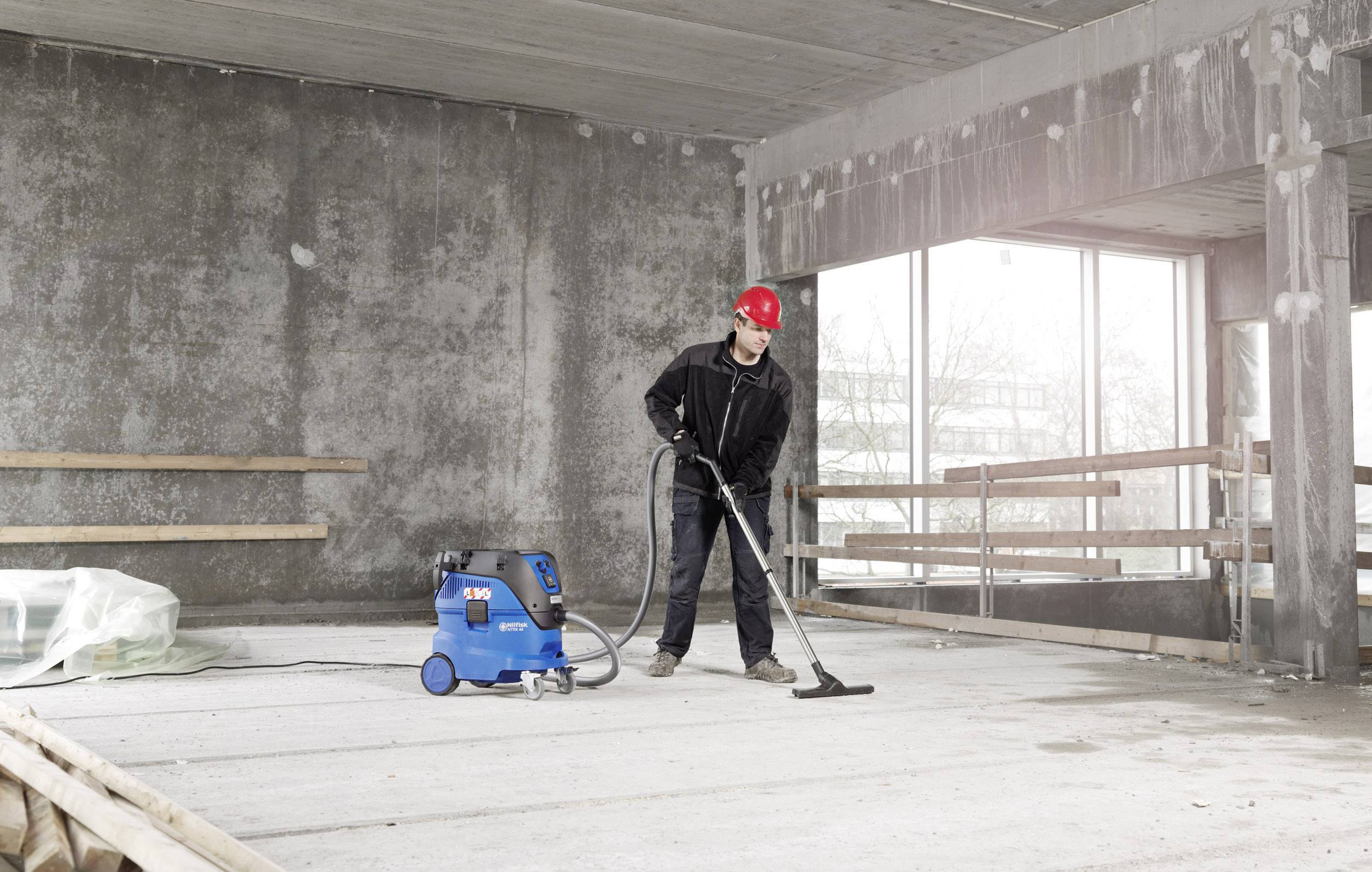 Ein Bauarbeiter mit rotem Helm saugt den Boden in einem unfertigen Raum mit einem blauen Industriestaubsauger.