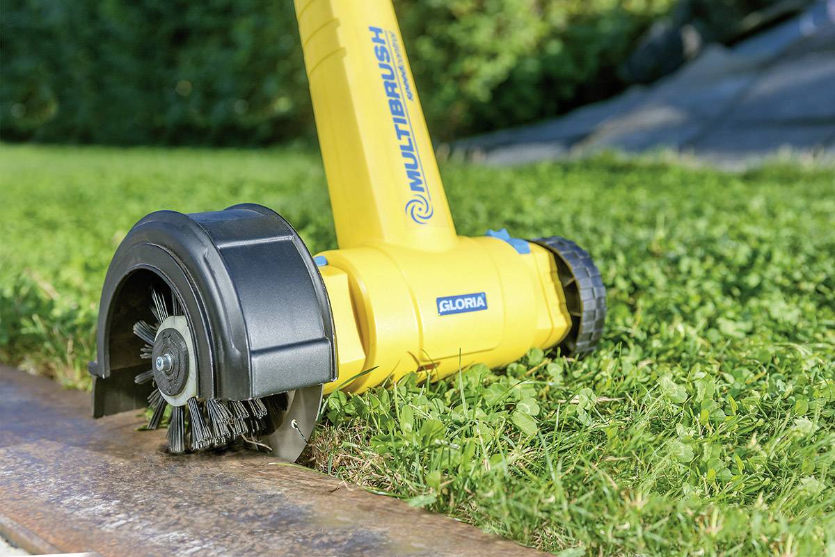 Ein gelbes Gartengerät mit Bürstenkopf reingt einen Steinrand im Garten, entfernt Moos und Unkraut von der Gehwegkante.