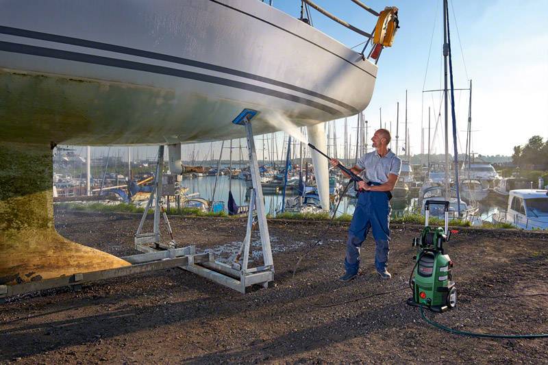 Eine Person reinigt ein Boot mit einem Hochdruckreiniger auf einem Bootsstellplatz nahe einem Hafen mit mehreren Yachten im Hintergrund.