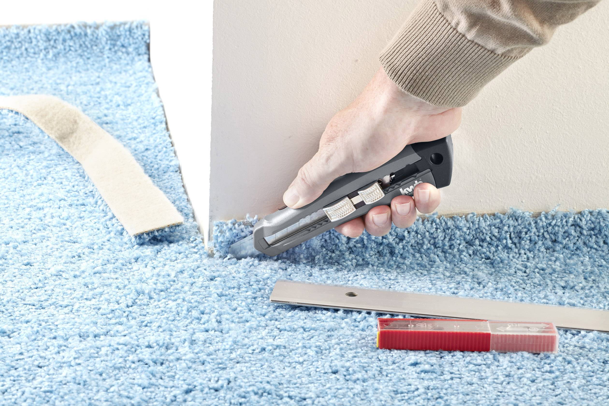Eine Person schneidet mit einem Teppichmesser blauem Teppichboden an einer Kante. Ein Metalllineal und ein Maßband liegen daneben.