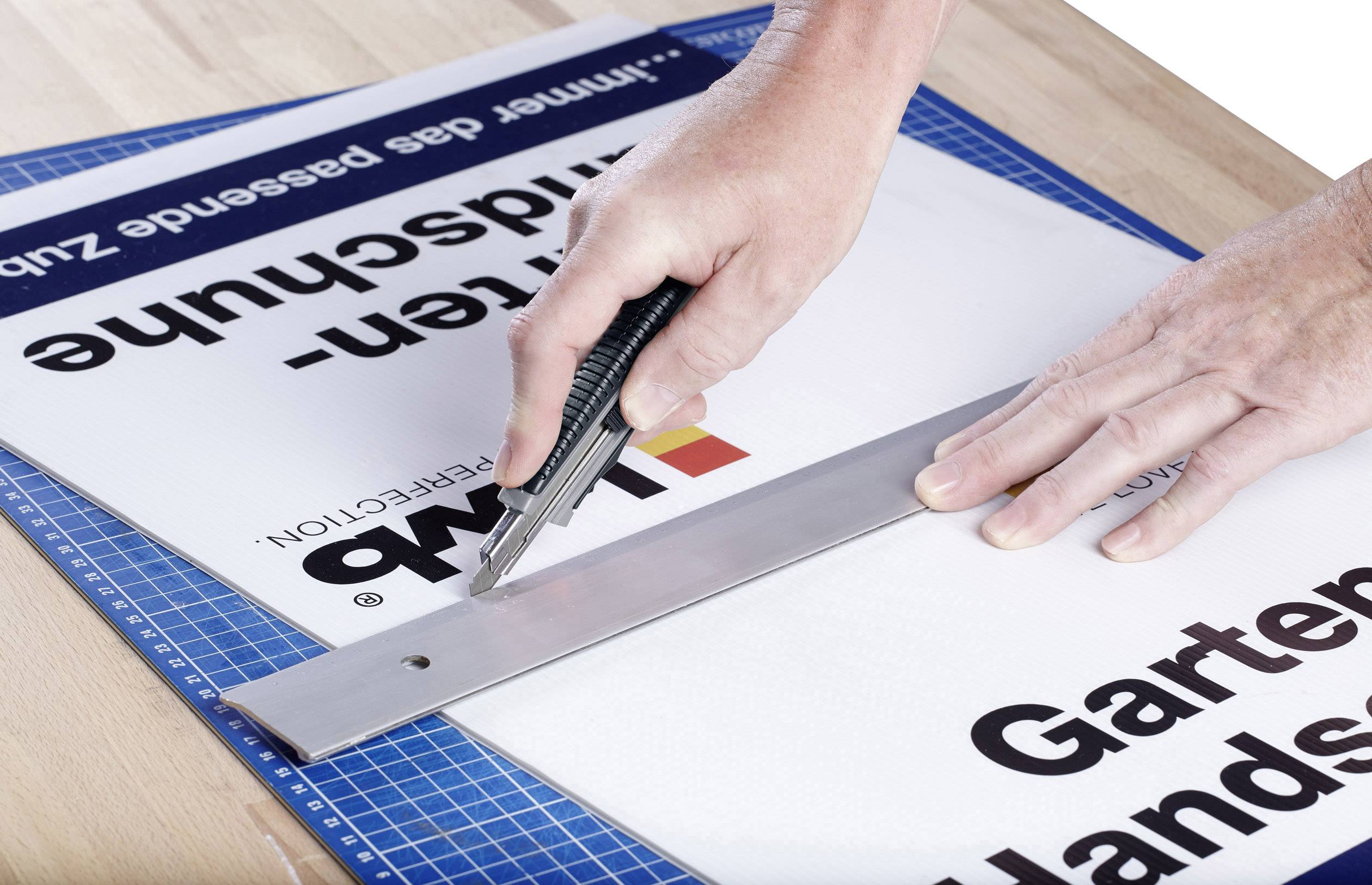 Eine Hand schneidet mit einem Cutter einen Schild entlang eines Lineals auf einem Schneidebrett. Auf dem Schild stehen unleserliche Textfragmente.