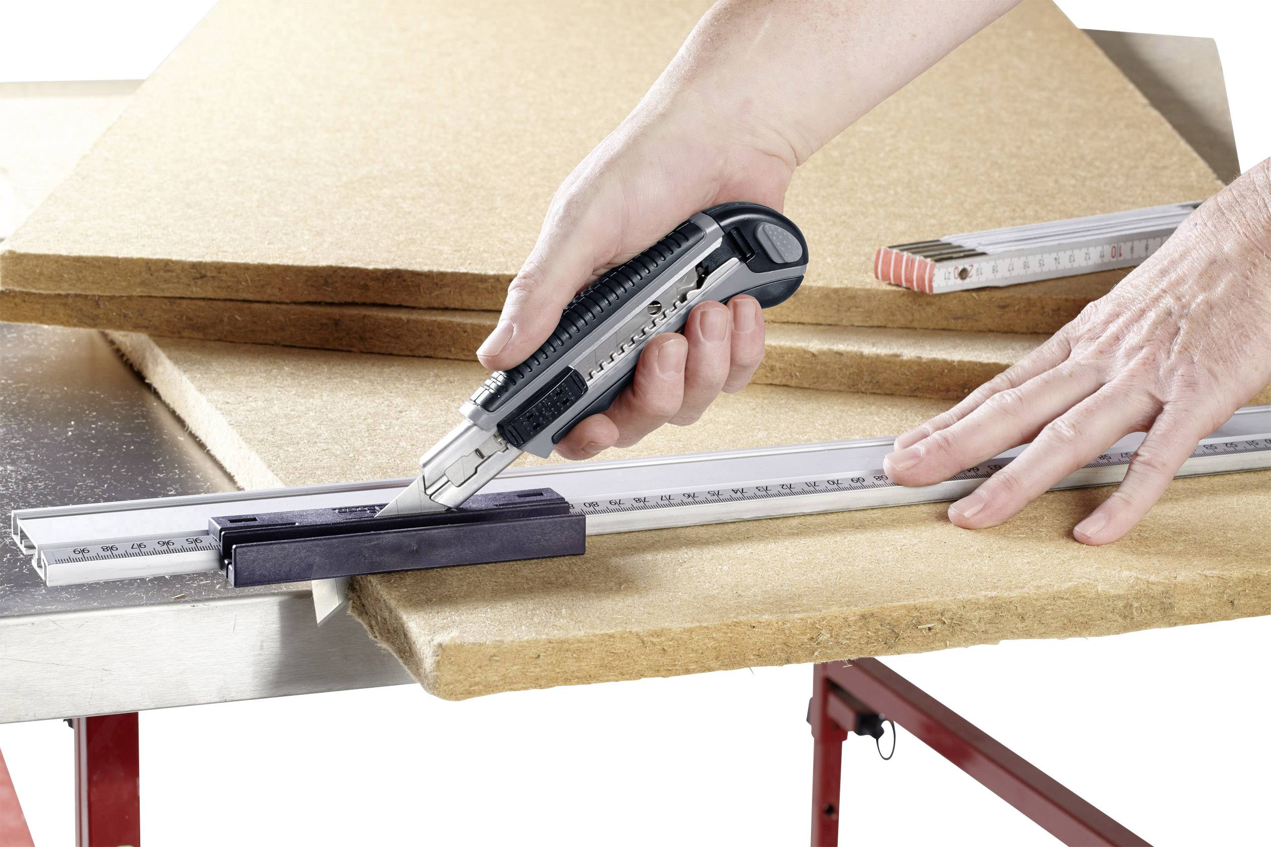 Eine Person schneidet eine Holzplatte mit einem Cutter-Messer entlang eines Metalllineals auf einem Arbeitstisch.
