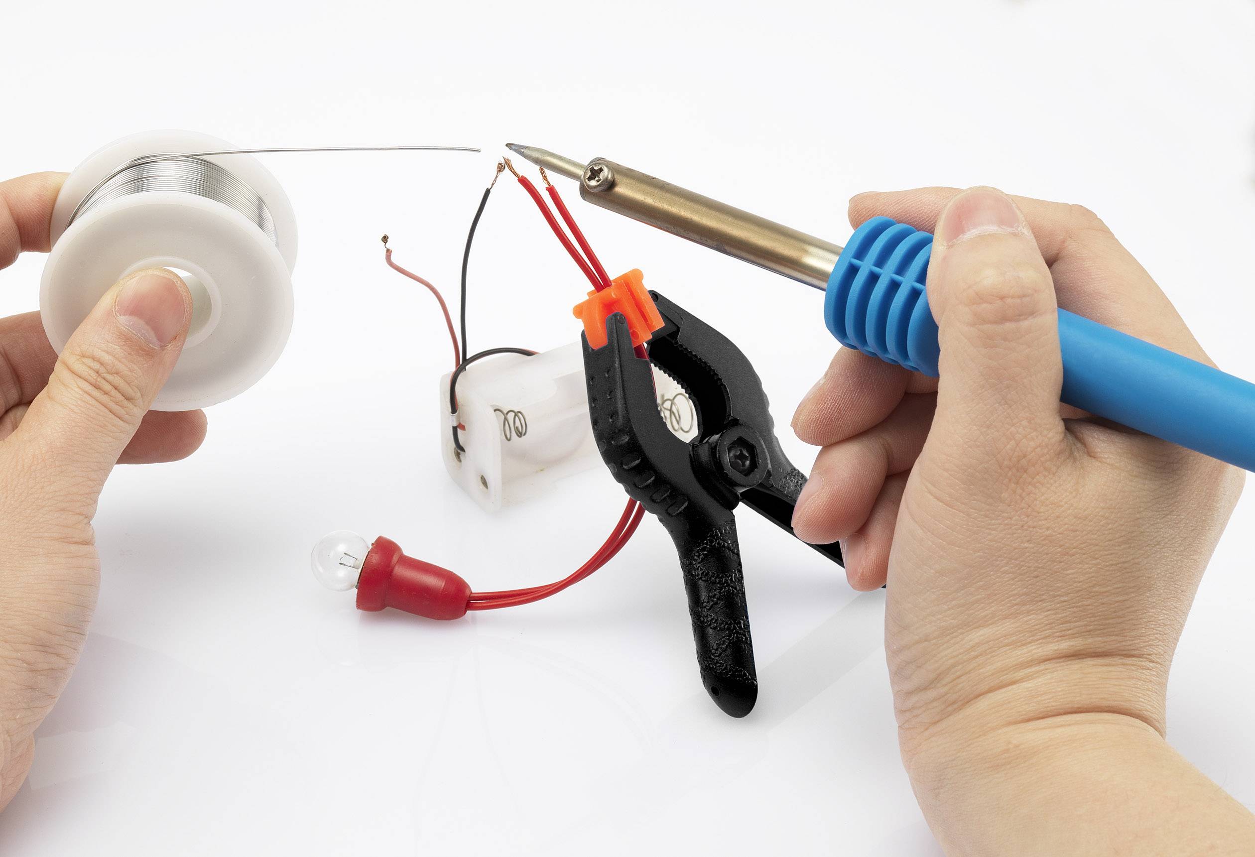 Eine Person lötet ein elektrisches Bauteil mit einem Lötkolben. Ein Kabel wird mit einer Klemme fixiert, während ein weiteres Kabel verlötet wird.