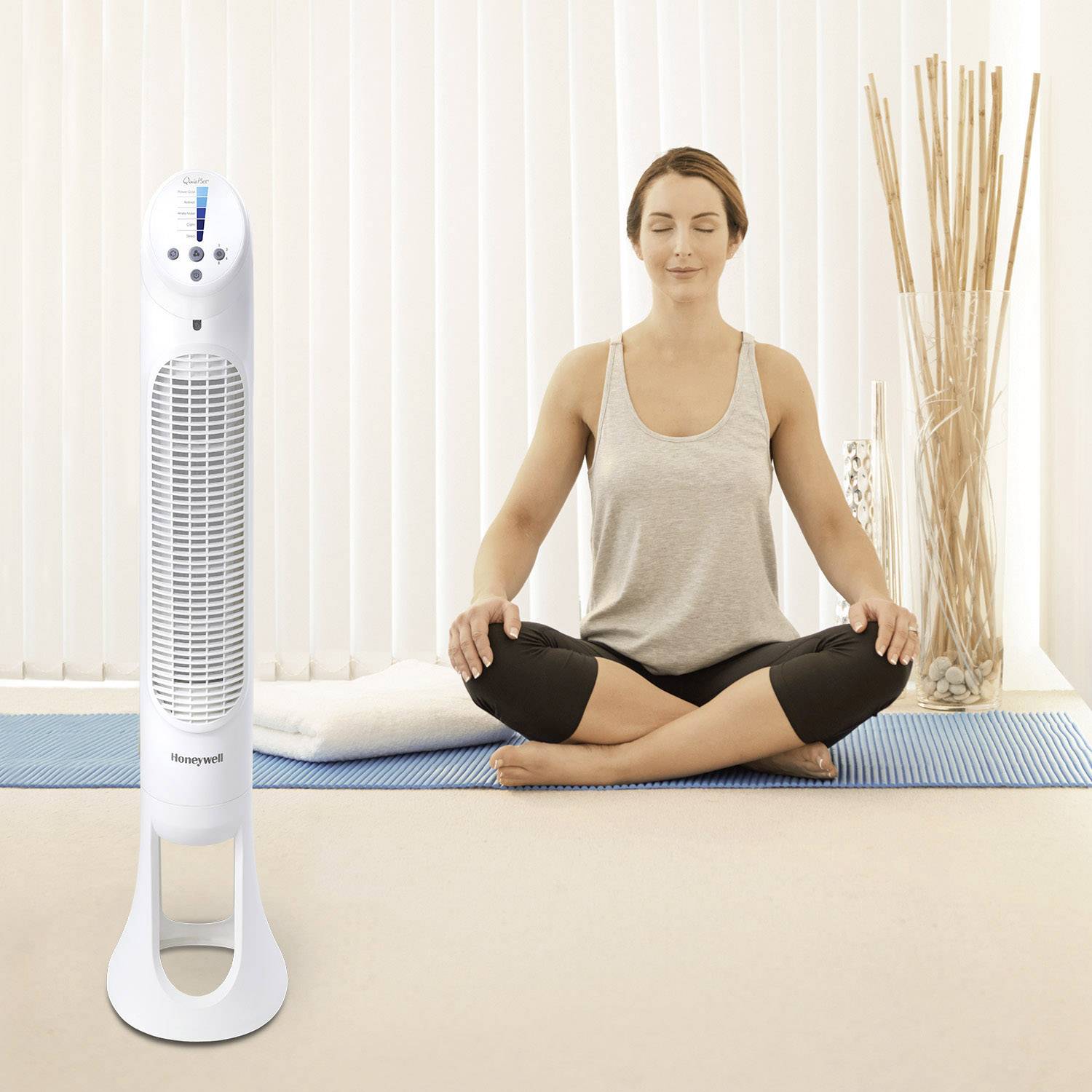 Frau meditiert im Wohnzimmer neben einem stehenden Ventilator vor einem Lamellenfenster. Sie sitzt im Schneidersitz auf einem Teppich.