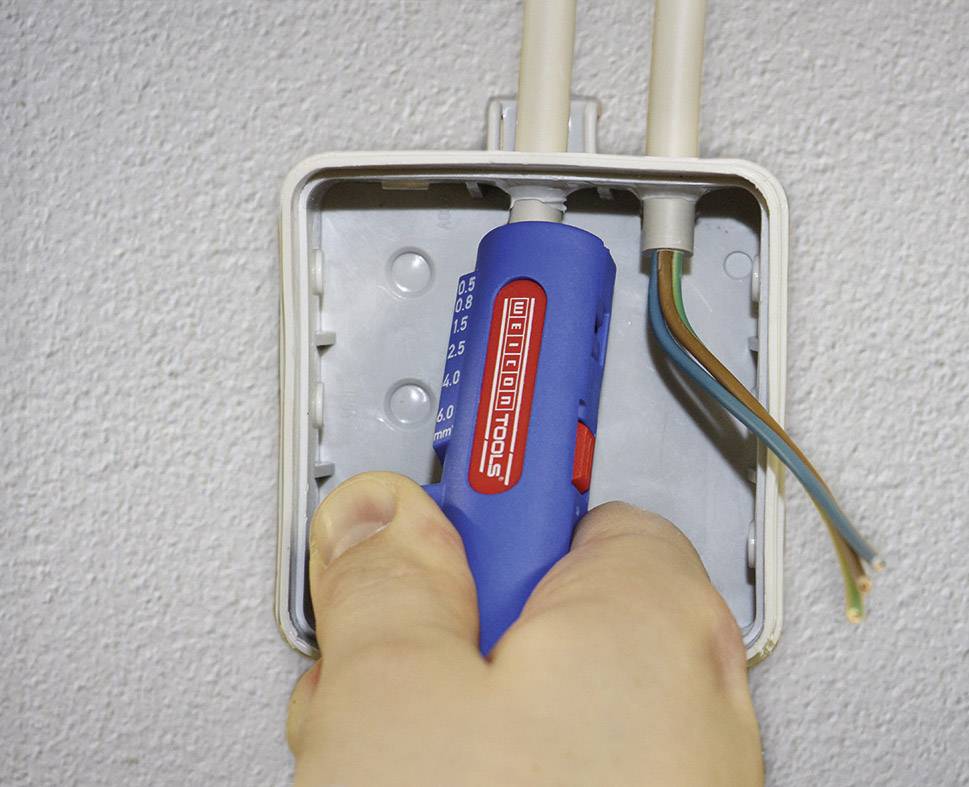 Hand hält einen Spannungsprüfer, der in eine offene Steckdose mit sichtbaren Drähten eingeführt wird. Sicherheit bei elektrischen Arbeiten.