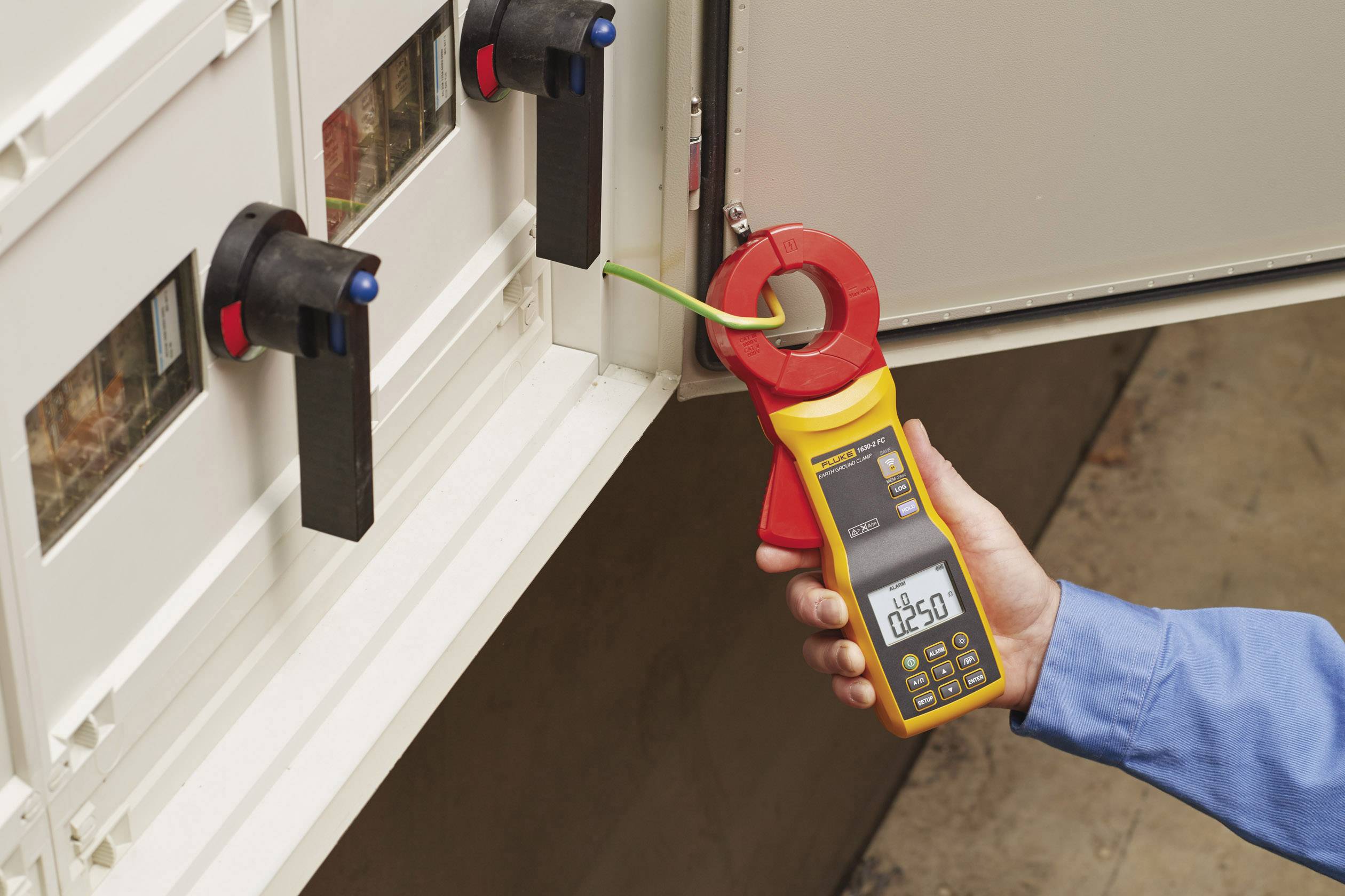 Eine Person misst den elektrischen Strom mit einer Digital-Zangenstromzange an einem grünen Kabel in einem Schaltschrank.