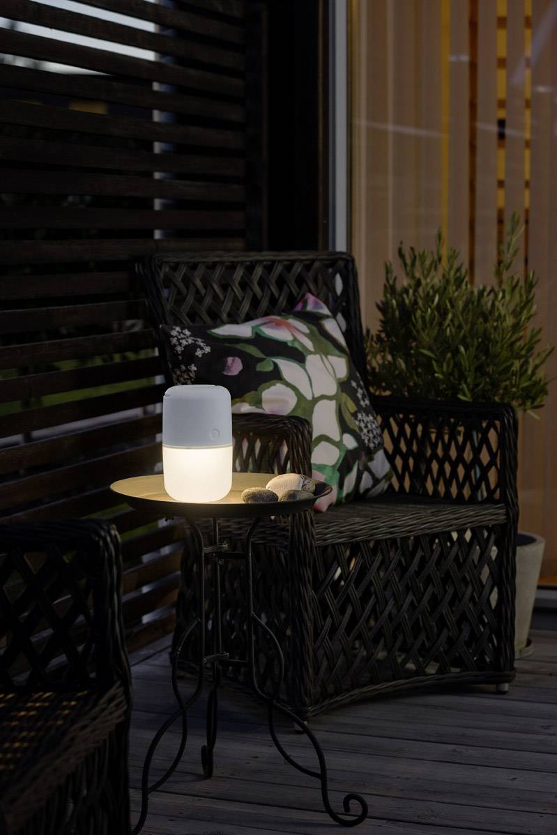 Ein beleuchteter Tisch auf einer Terrasse mit Korbsessel und Pflanze. Auf dem Tisch steht eine leuchtende Lampe neben einer kleinen Rolle.